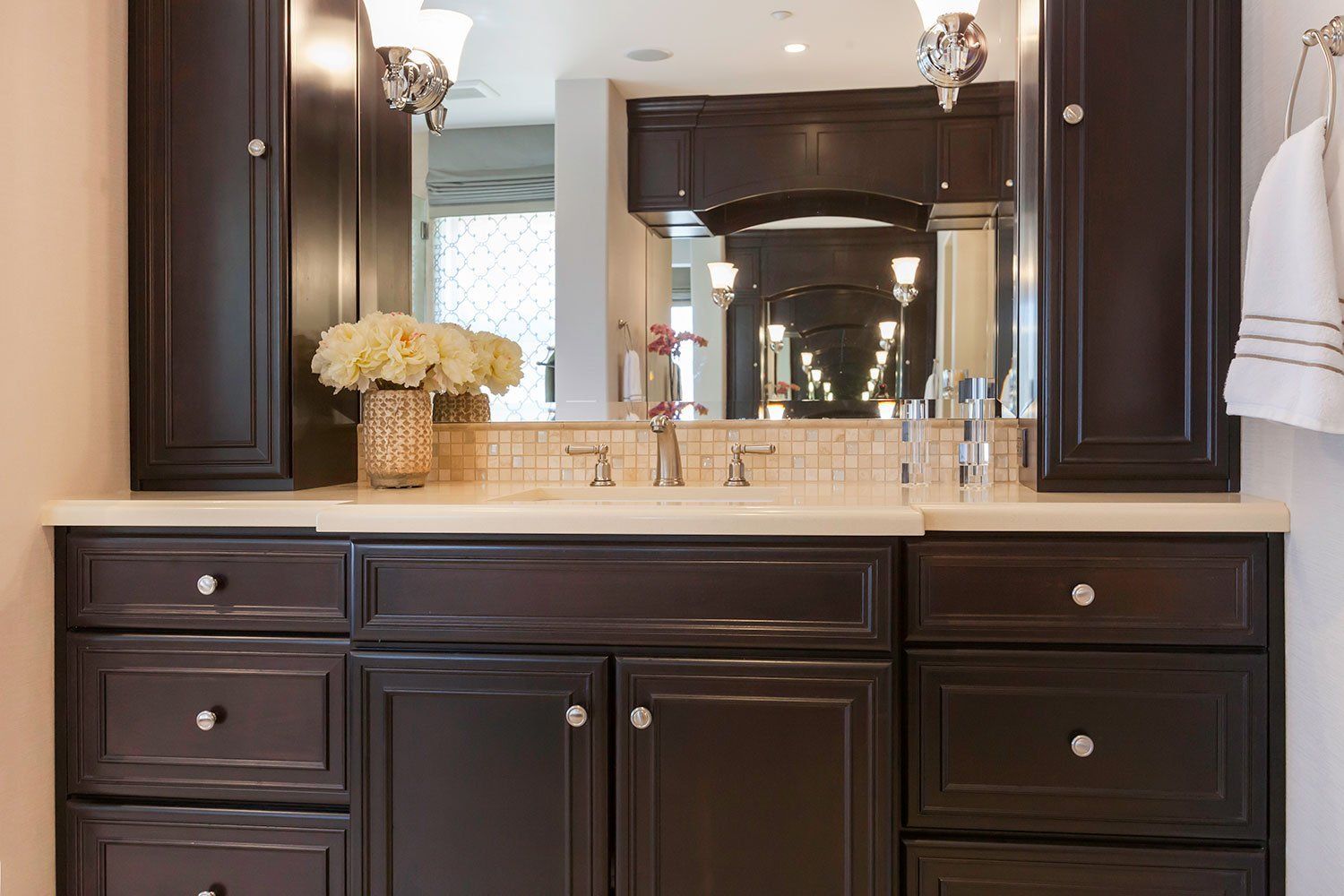 Bathroom vanity with dark wood cabinets, beige countertop, large mirror, and white flowers