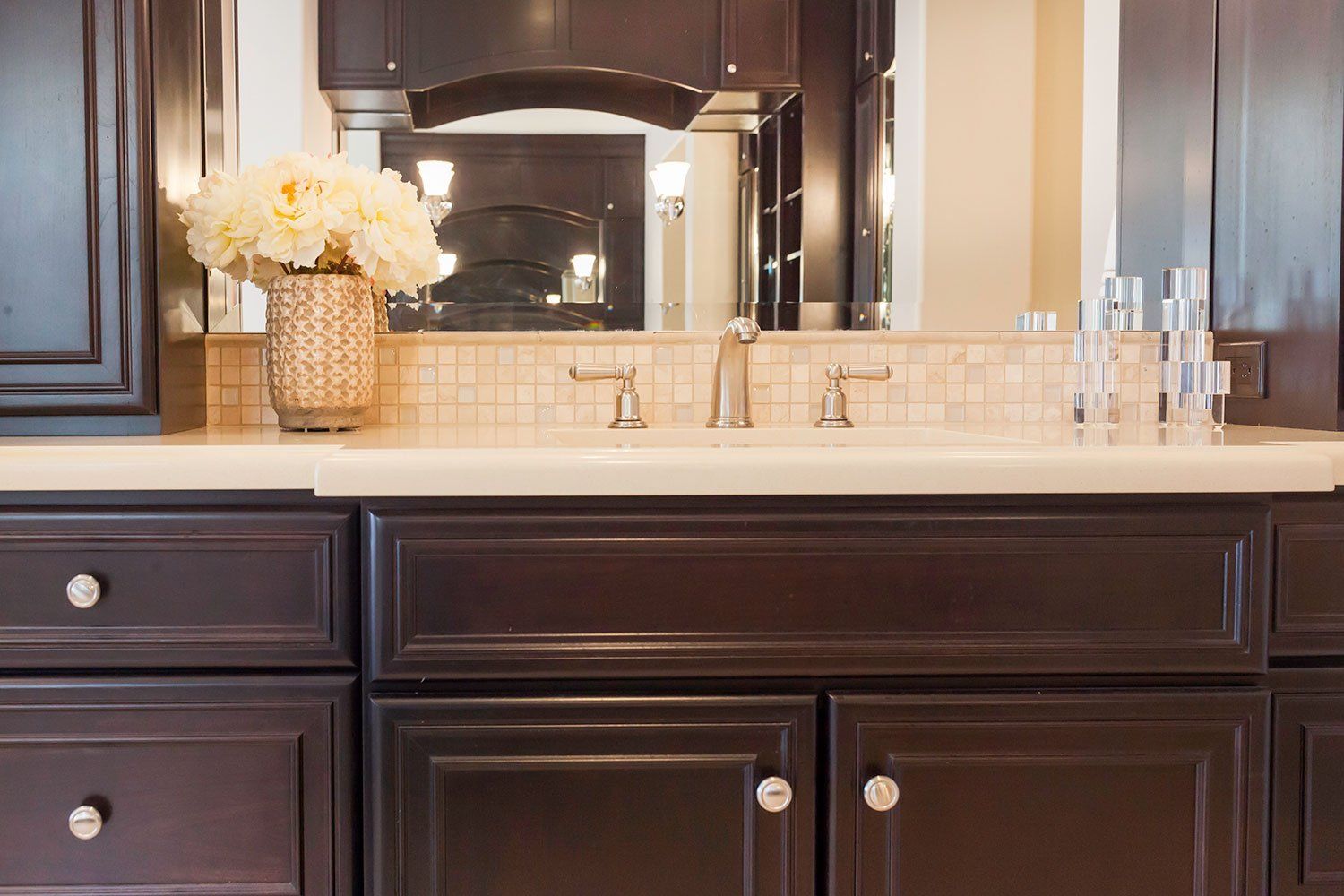 Dark wood bathroom vanity with white countertop, mosaic backsplash, and vase of white flowers