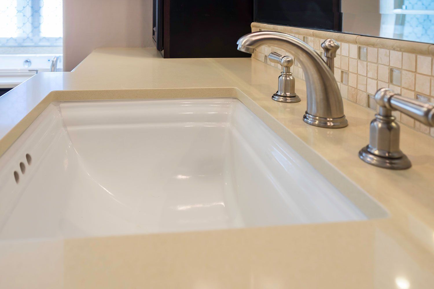 White kitchen sink with beige countertop and chrome faucet in a tiled kitchen.