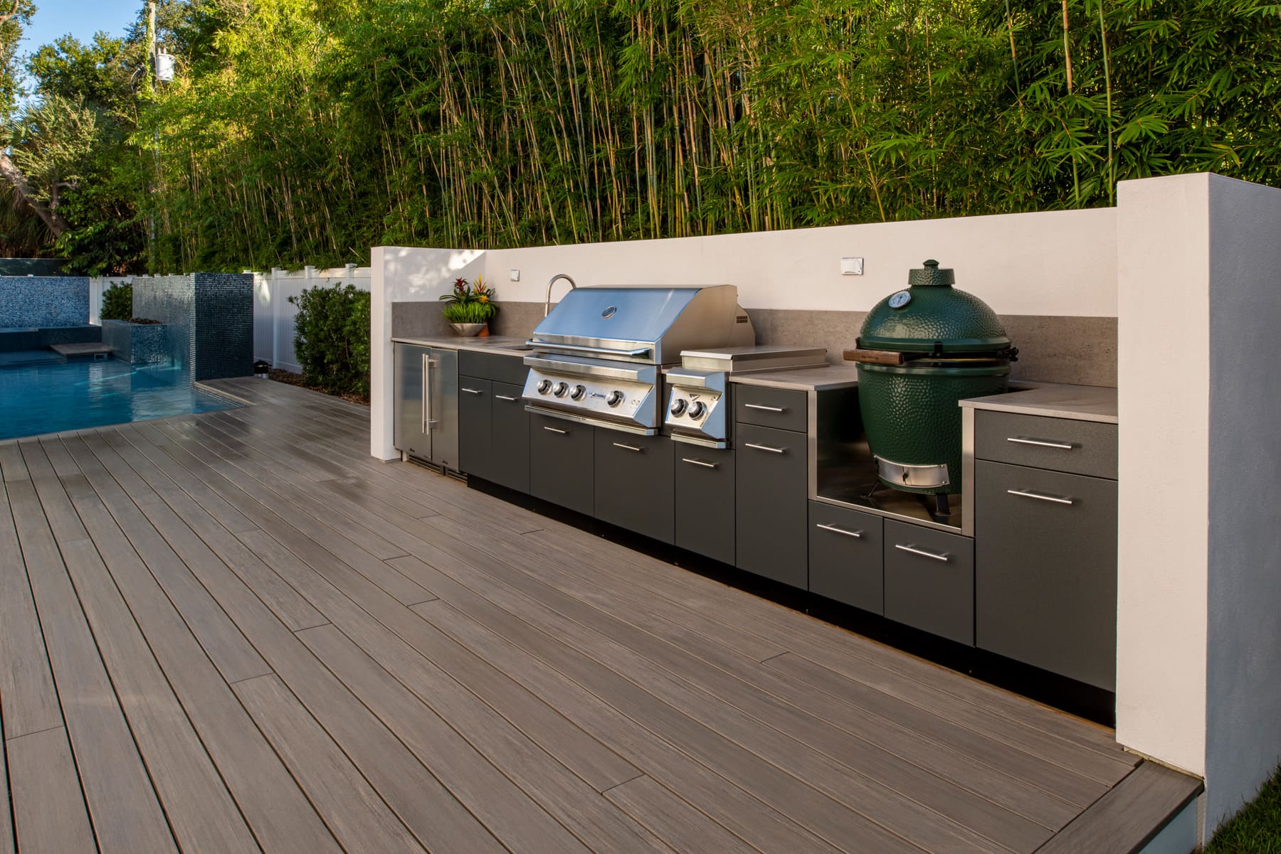 Outdoor patio with built-in grill and sink beside a pool, set against a white wall and greenery