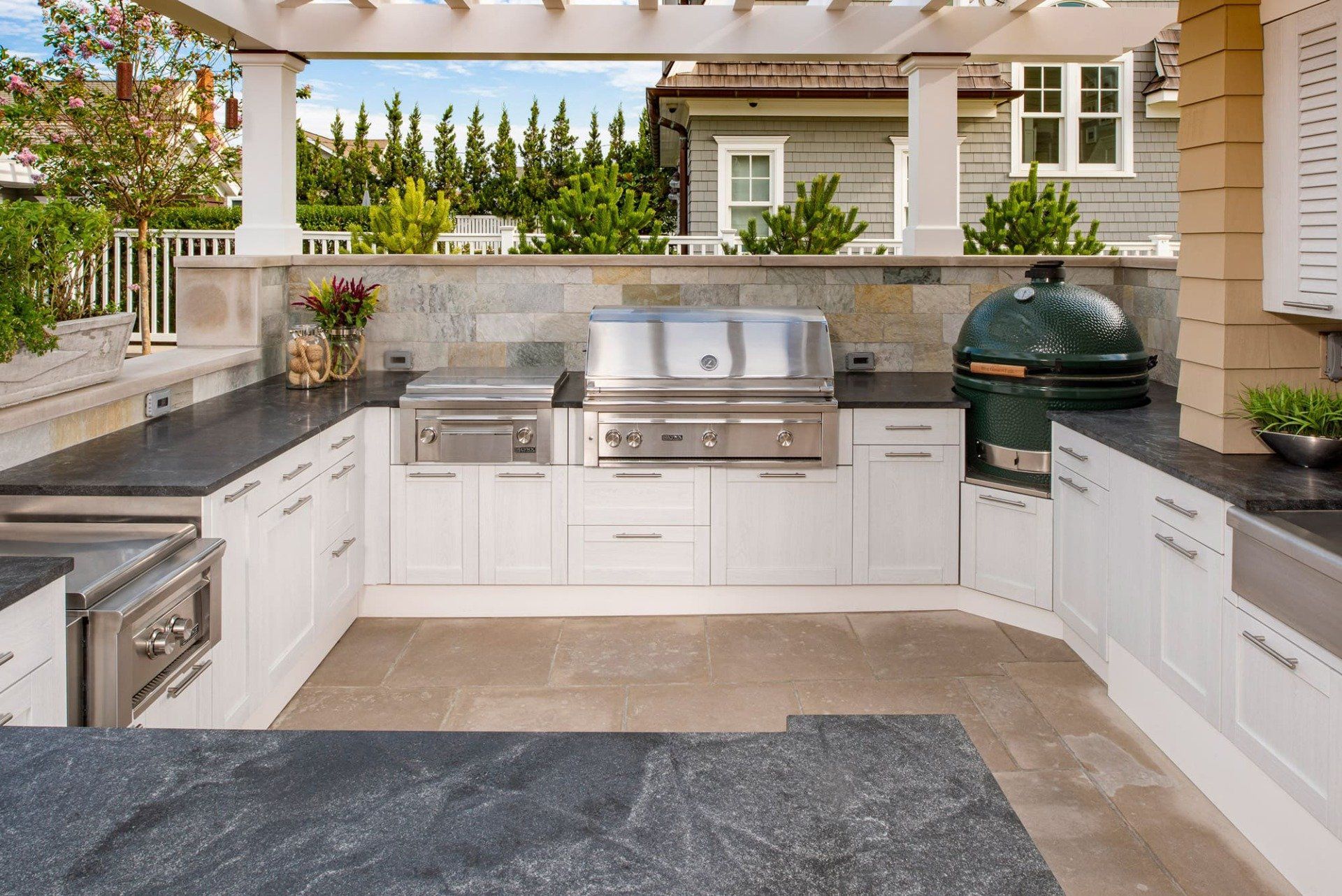Outdoor kitchen with white cabinets, stainless grill, black smoker, and stone countertop under a pergola