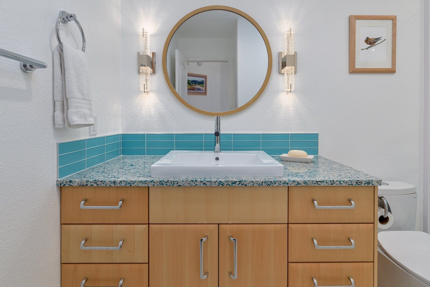 Bathroom vanity with gold round mirror, turquoise tile backsplash, wooden cabinets, and white countertop sink