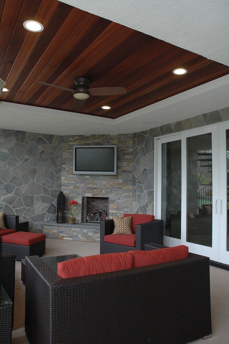 Covered patio lounge with wicker seating, stone fireplace, wall TV, and wooden ceiling with recessed lights