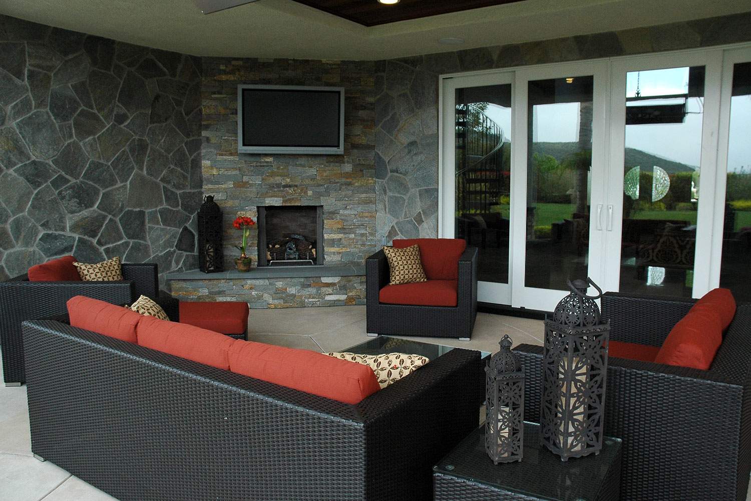 Covered patio with wicker seating, red cushions, stone fireplace, and glass doors overlooking a yard