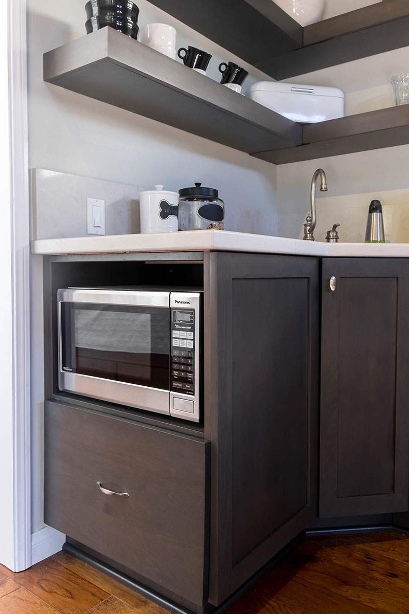 Compact kitchen nook with a built-in microwave, sink, and dark wood cabinets beneath floating shelves.