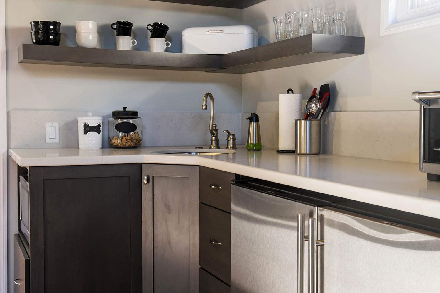 Modern kitchen corner with dark cabinets, white countertops, and shelves holding jars, mugs, and utensils