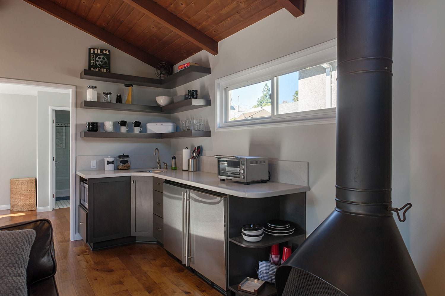 Small kitchen with white cabinets, open shelves, wood ceiling, and black wood stove by a window.