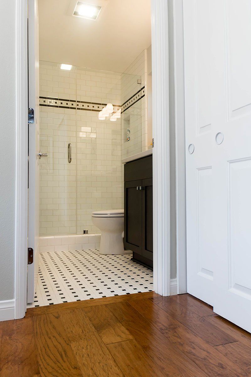 Open bathroom doorway with white tile, patterned floor, and dark vanity visible from hardwood hallway