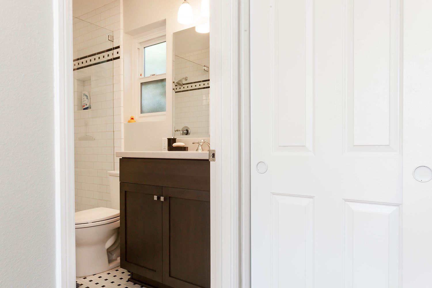 Small bathroom with white tile, dark vanity, toilet, mirror, and shower curtain partially visible.