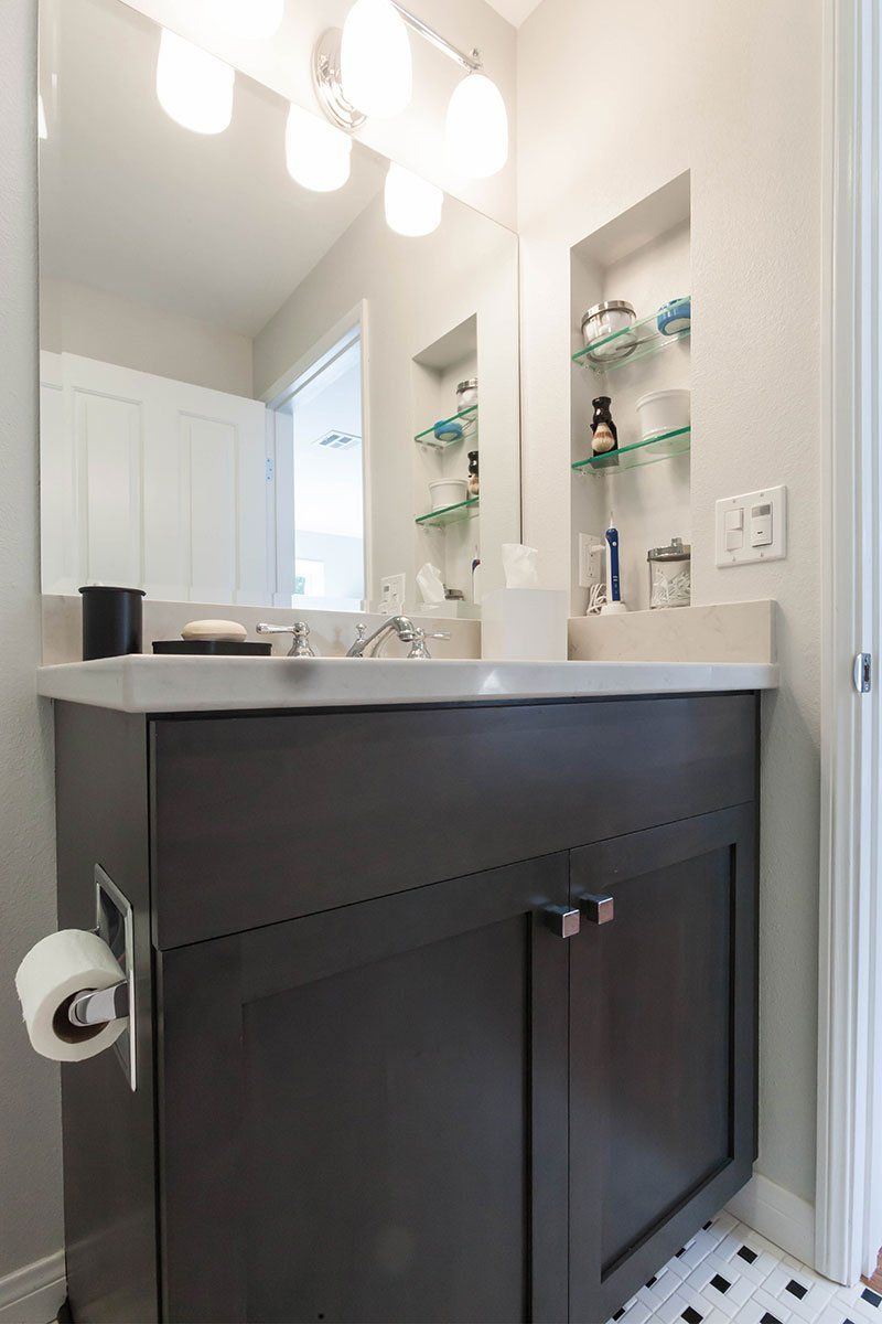 Narrow bathroom vanity with dark cabinet, white sink, mirror, and shelves beside a white door.