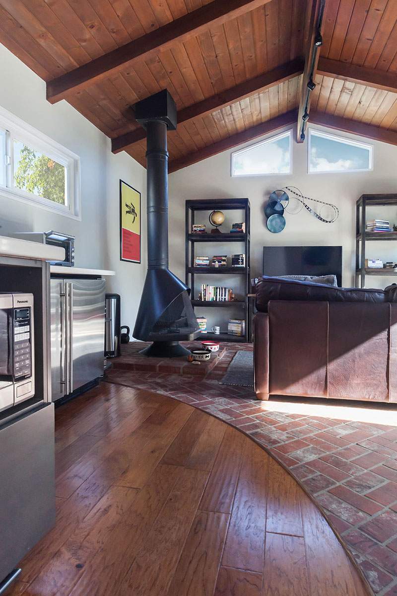 Cozy cabin living room with wood ceiling, black stove, bookshelves, and curved hardwood floor