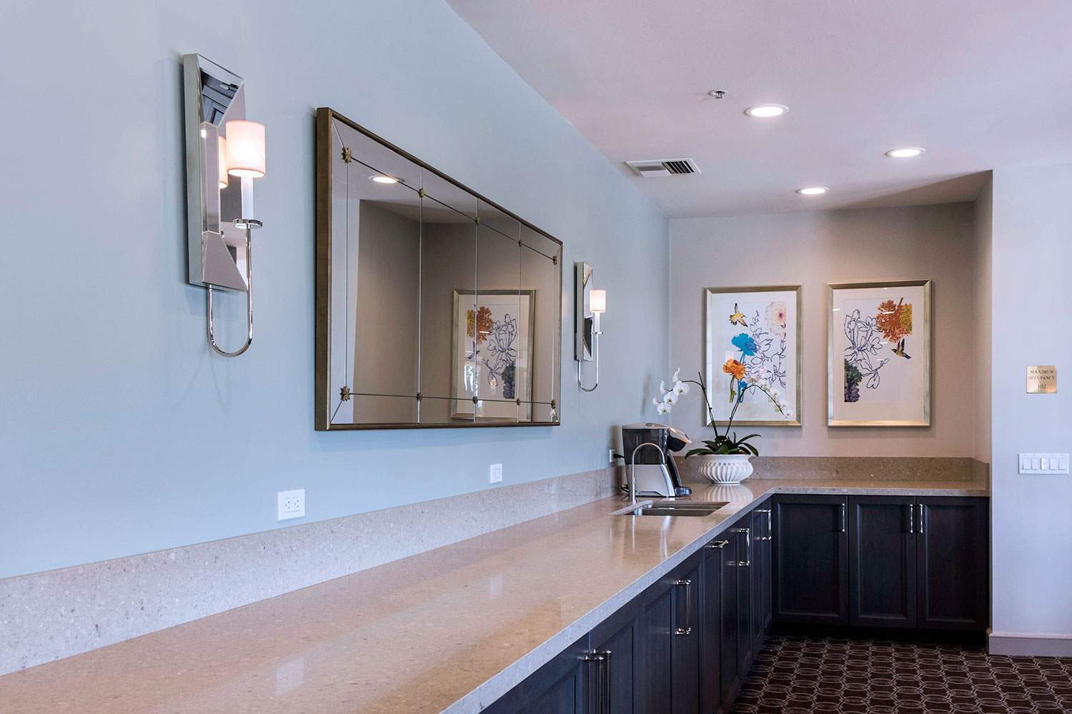 Empty hotel bathroom vanity with mirror, light blue walls, dark cabinets, and framed artwork.