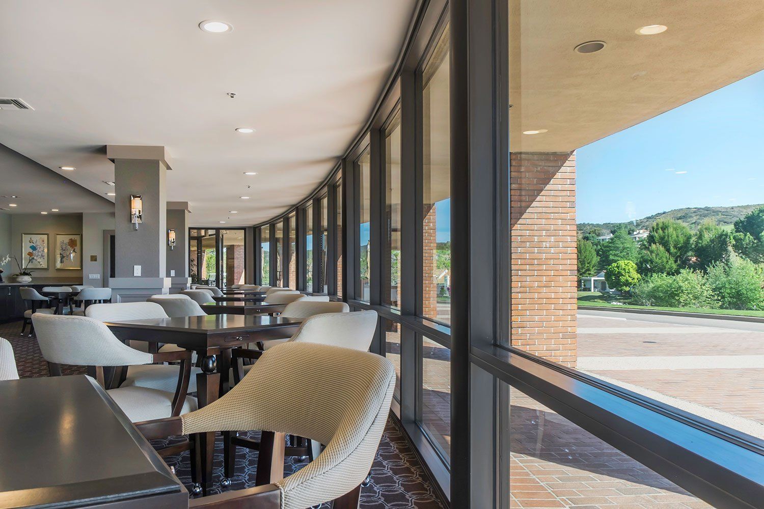 Empty café seating area beside large windows overlooking a sunny road and green trees