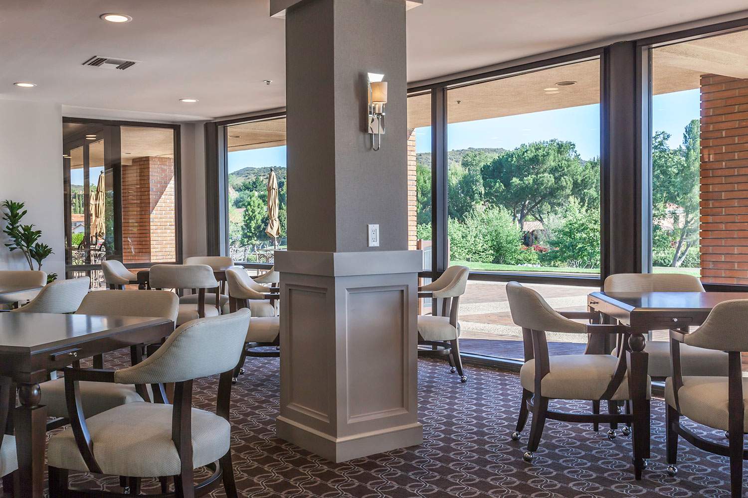Bright dining room with empty tables, upholstered chairs, and large windows overlooking greenery