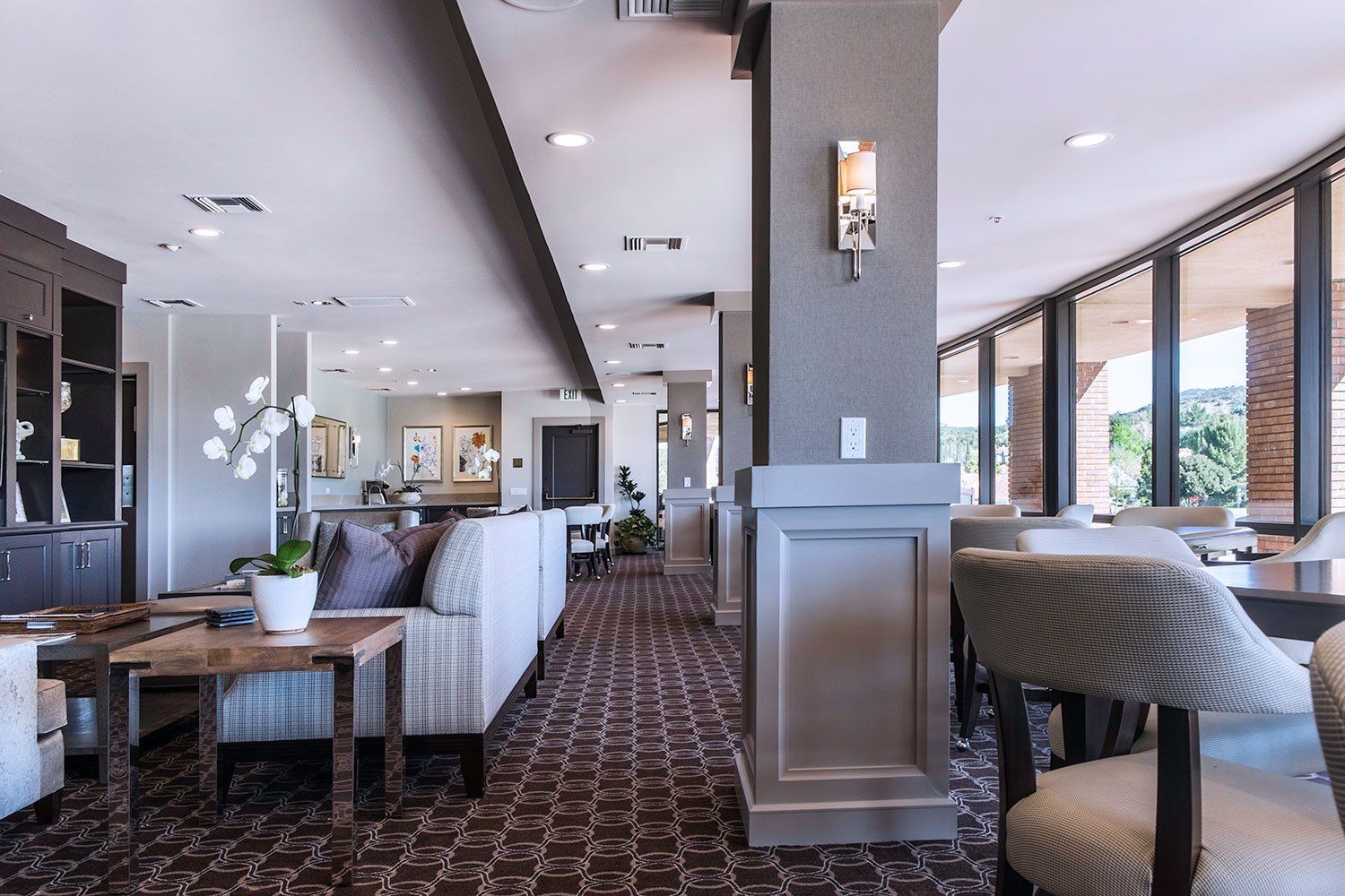 Modern hotel lounge with patterned carpet, white seating, large windows, and softly lit ceiling fixtures