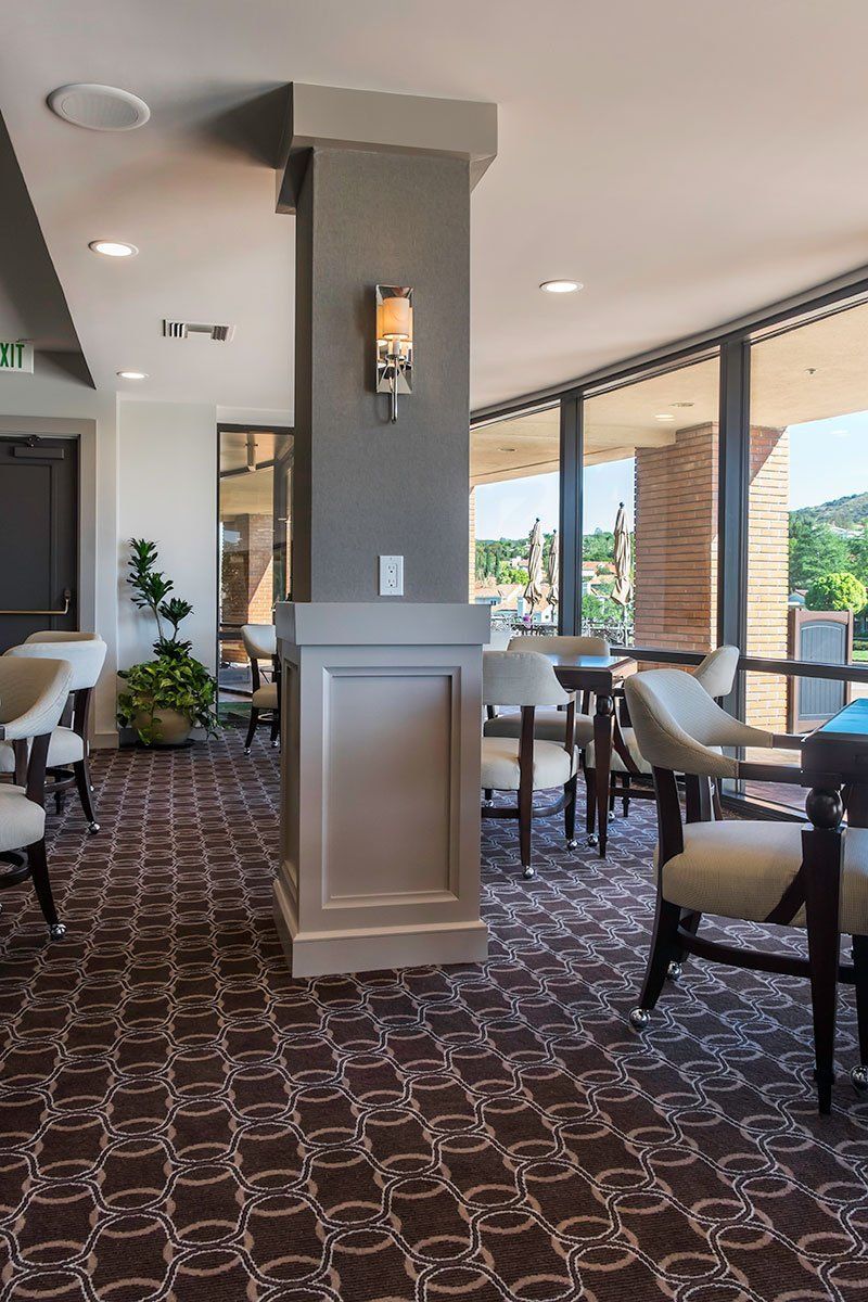 Upscale restaurant dining area with patterned carpet, gray columns, and large windows overlooking greenery