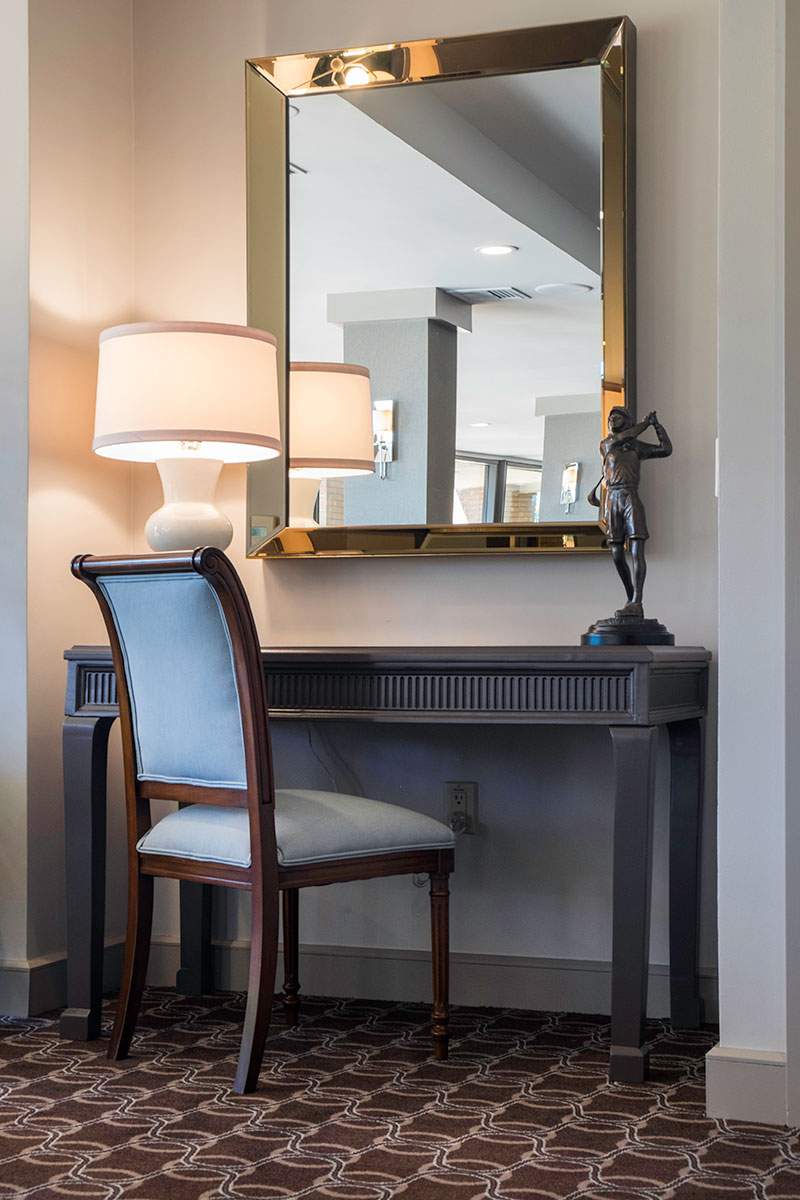 Elegant desk nook with lamp, mirror, chair, and decorative statue against a wall.