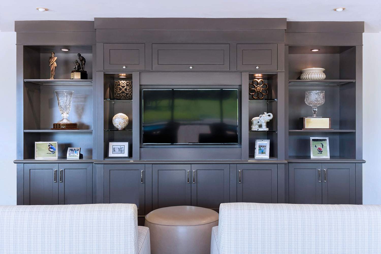 Gray built-in living room wall unit with TV, shelves, and decor above a couch and ottoman.