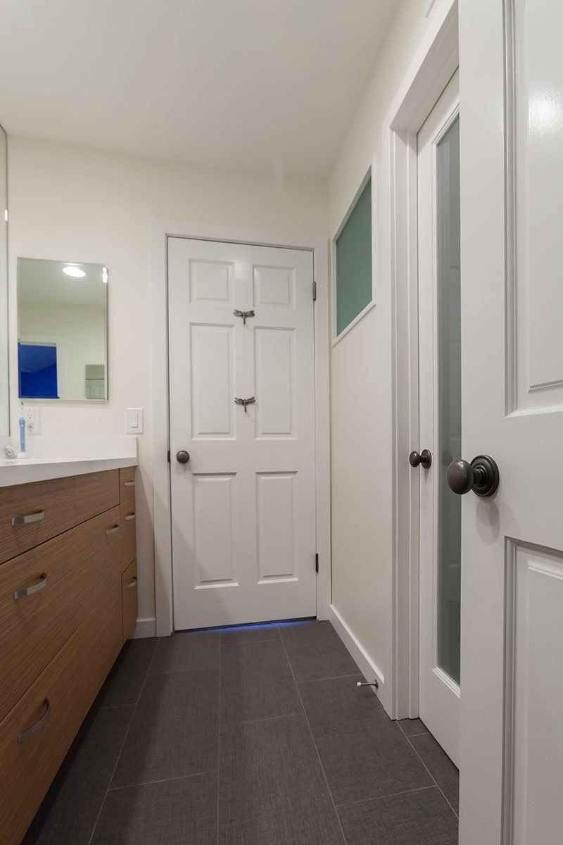 Narrow bathroom hallway with white doors, dark floor, and a wood vanity with mirror on the left.