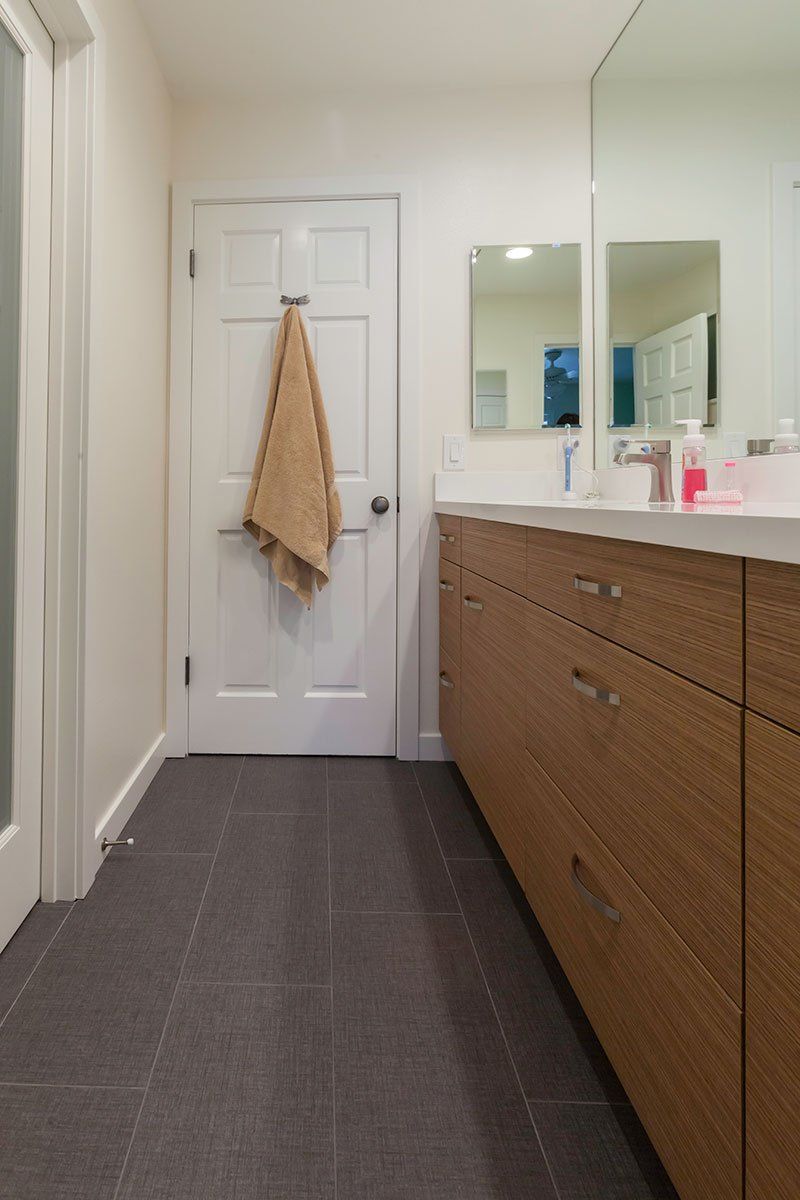 Narrow bathroom with white walls, gray tile floor, wood vanity, and a towel hanging on the door
