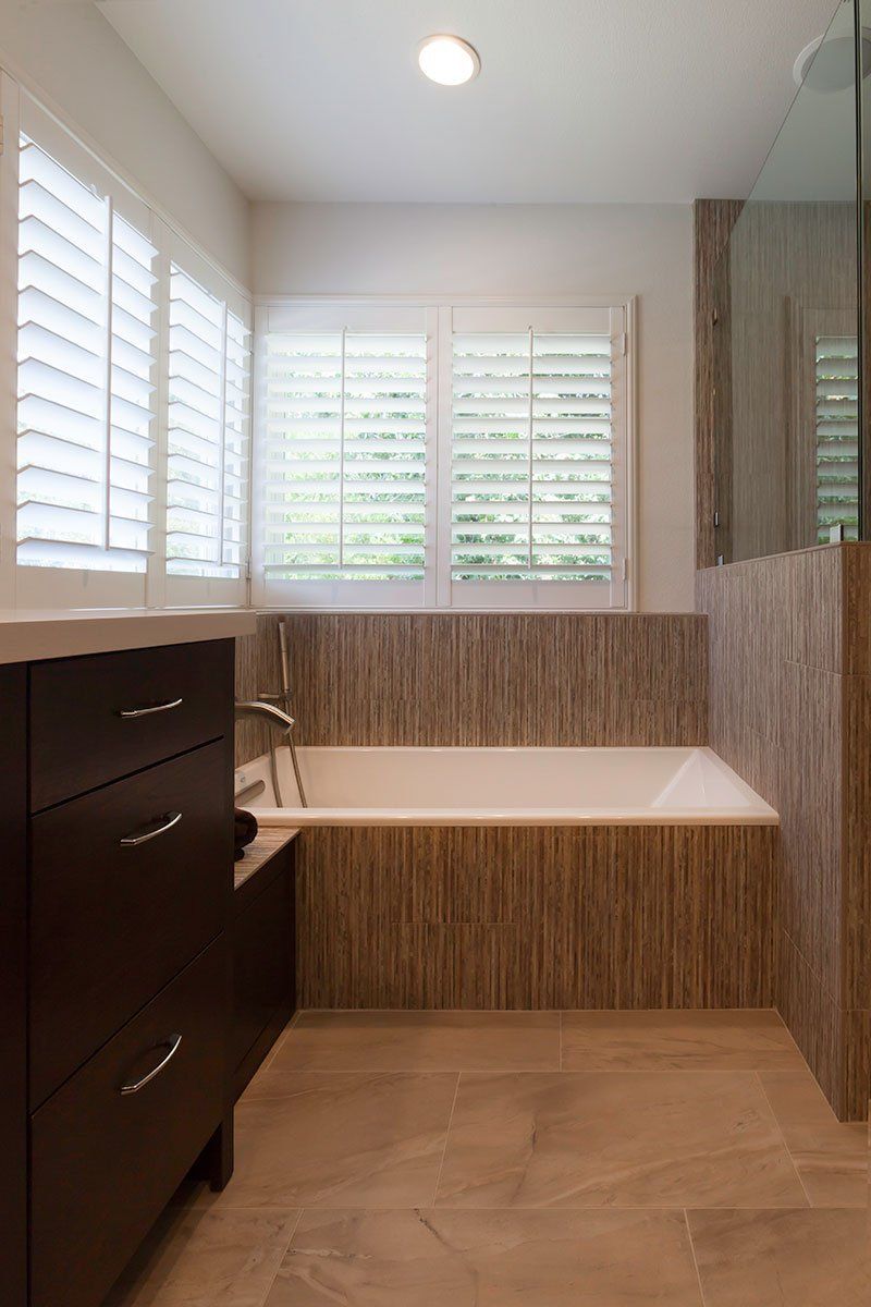 Modern bathroom with a built-in white tub, wood-look tile, dark vanity, and shuttered windows
