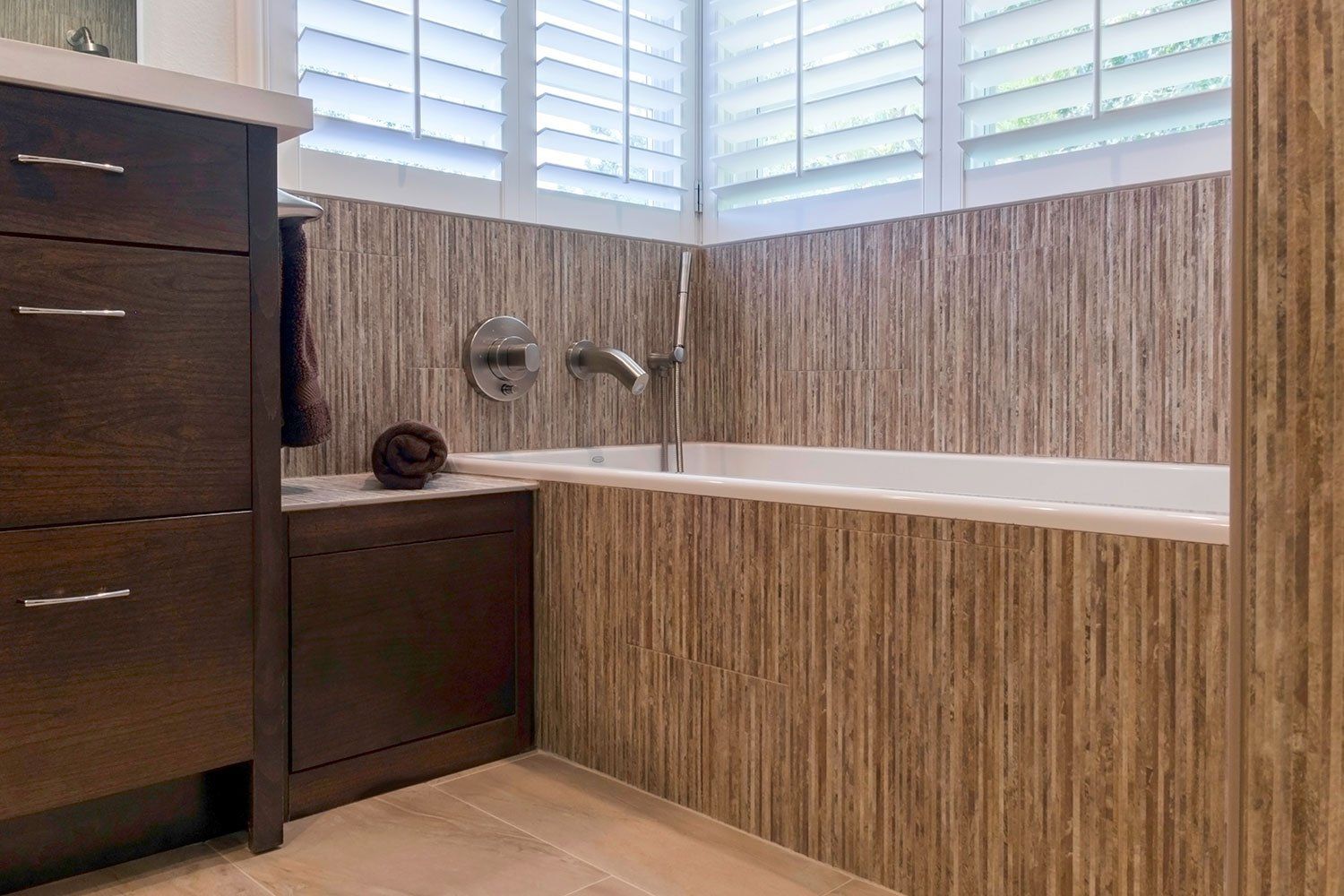 Modern bathroom with wood vanity, white bathtub, and horizontal striped wall tiles near a window