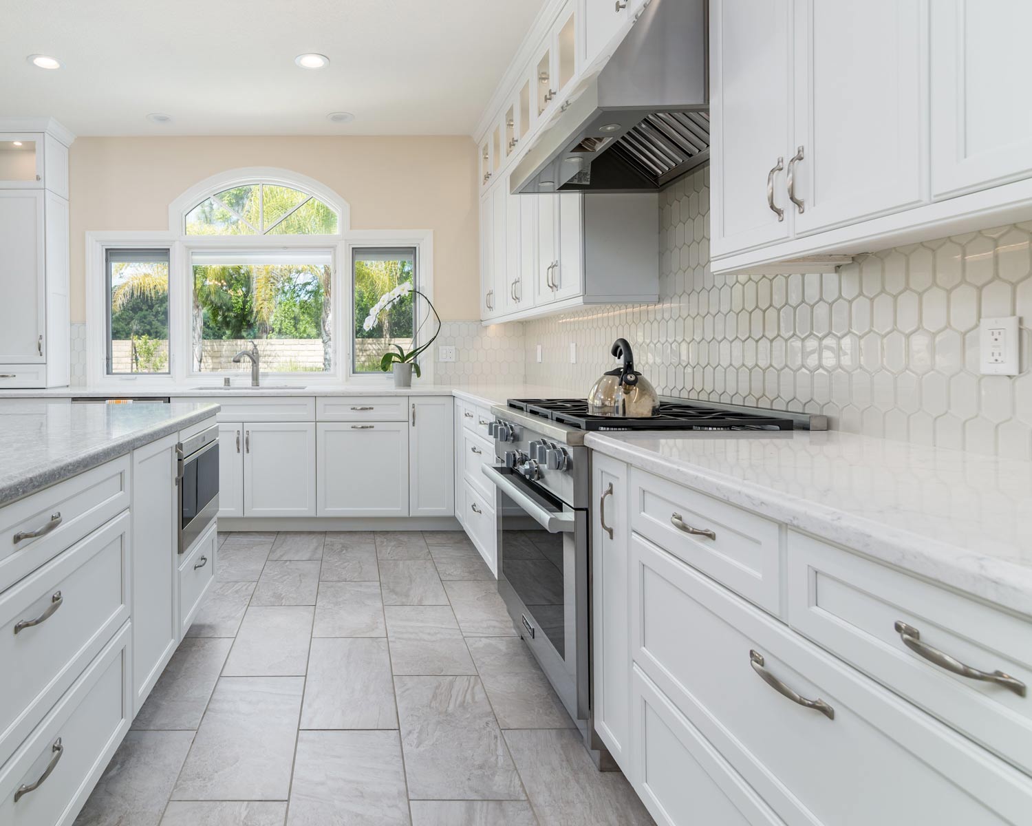 Bright white kitchen with marble island, stainless stove, tiled backsplash, and large window.