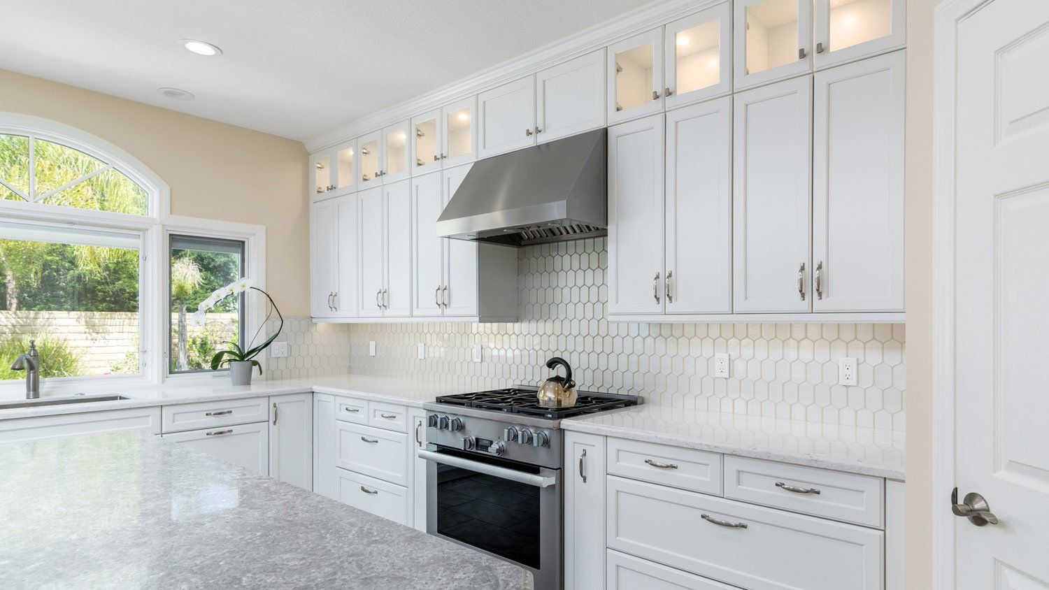 Bright white modern kitchen with cabinets, granite island, stainless stove, and large window over sink