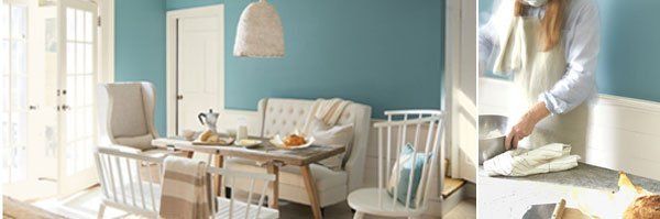Bright living room with a sofa, dining table, and a person baking in a sunlit kitchen.