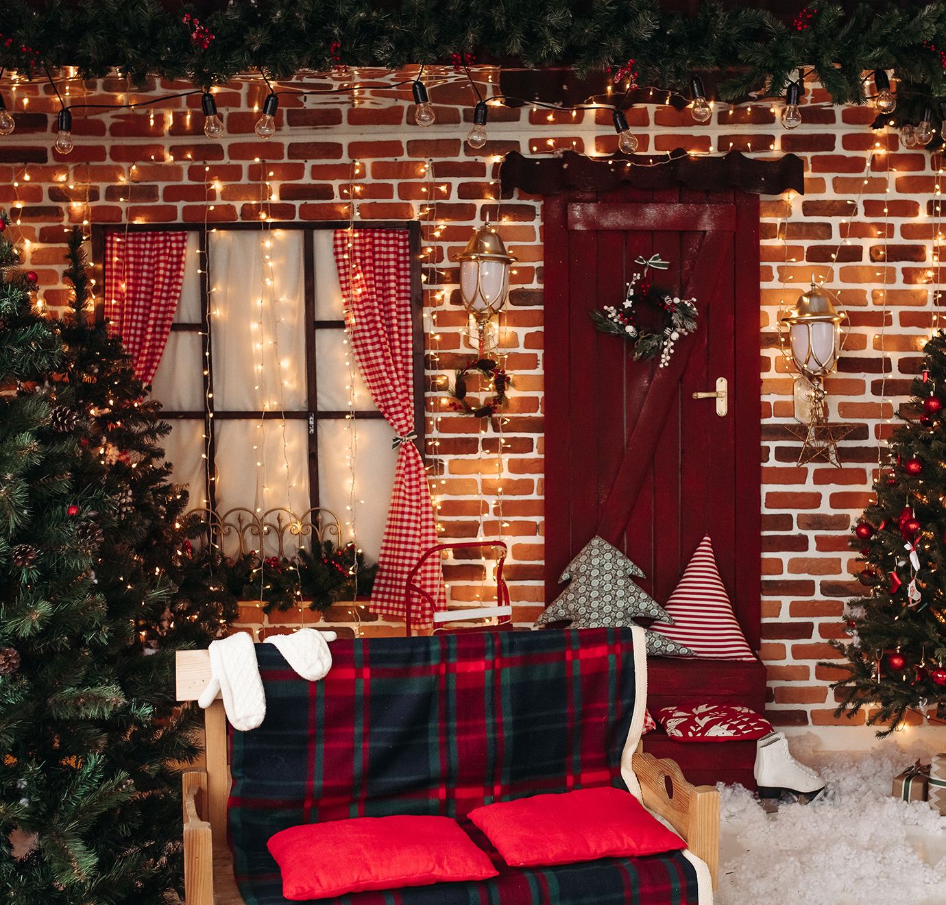 Brick Christmas scene with a red door, window, and plaid blanket. Christmas trees and lights add to the holiday feel.