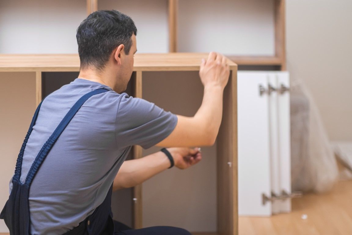 Person assembling a wooden cabinet in a room, reaching into an open shelf beside hanging fabric samples