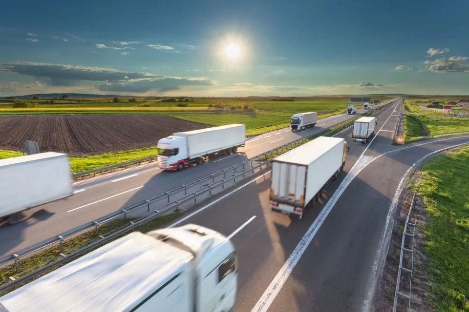 Trucks traveling on a highway beside green fields under a bright sky at sunset
