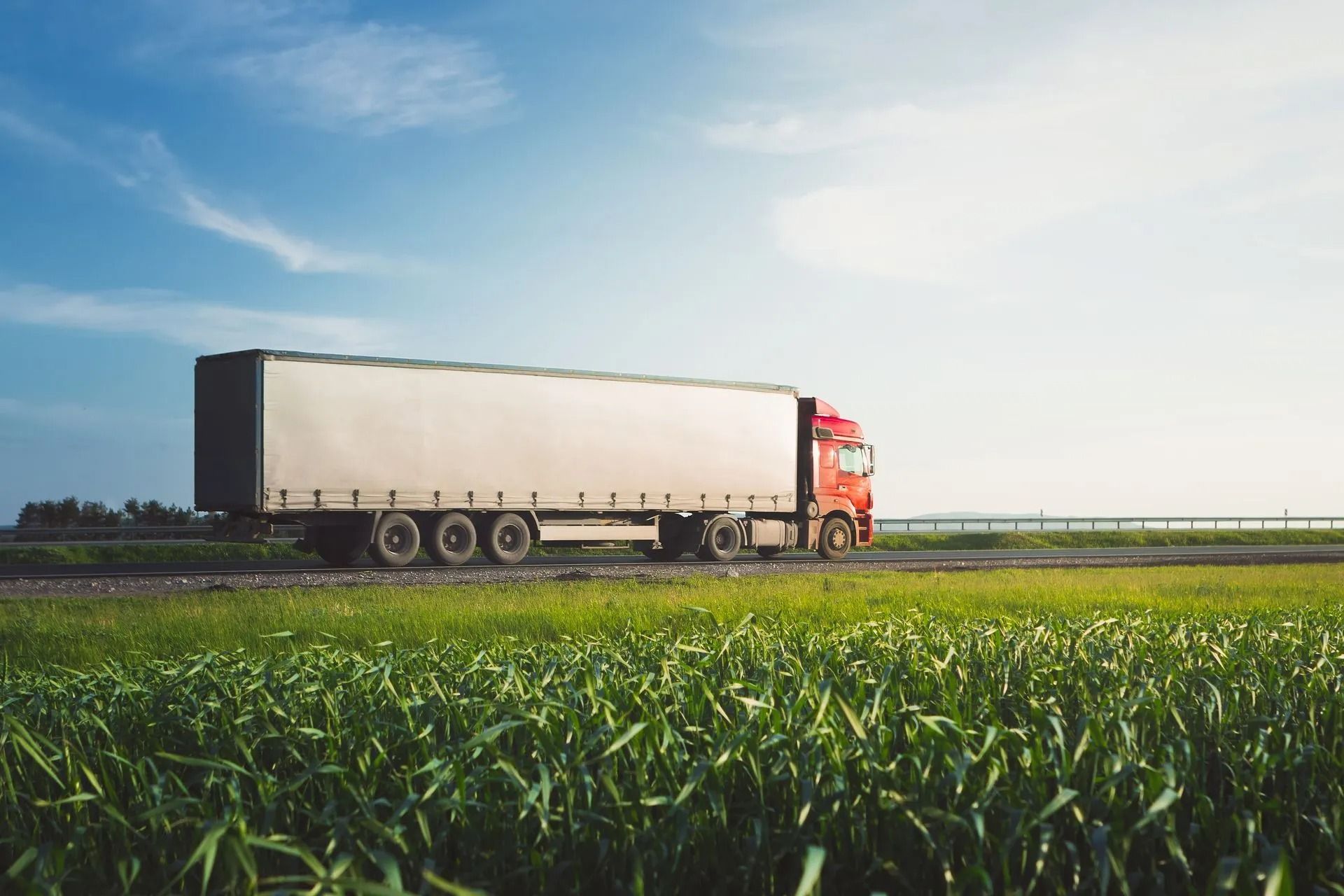 Red semi-truck hauling a white trailer driving beside a green field under a blue sky.