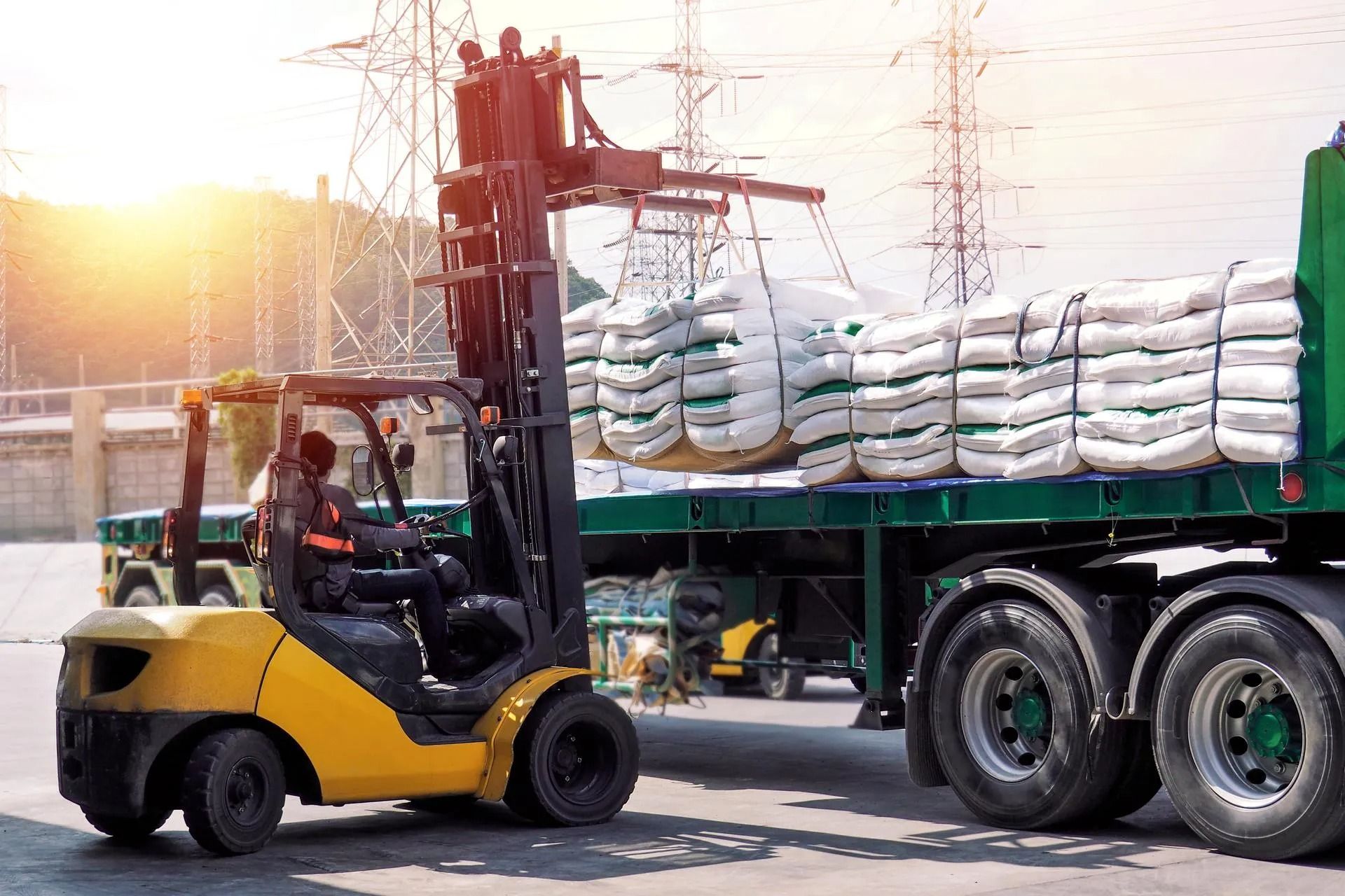 Yellow forklift loading white sacks onto a flatbed truck in an industrial yard at sunrise