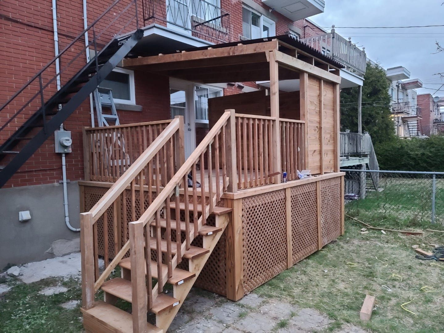 Rampe d'accès et escalier en bois donnant sur une cour arrière, attenants à une maison en briques, avec rambardes et plinthes en treillis.