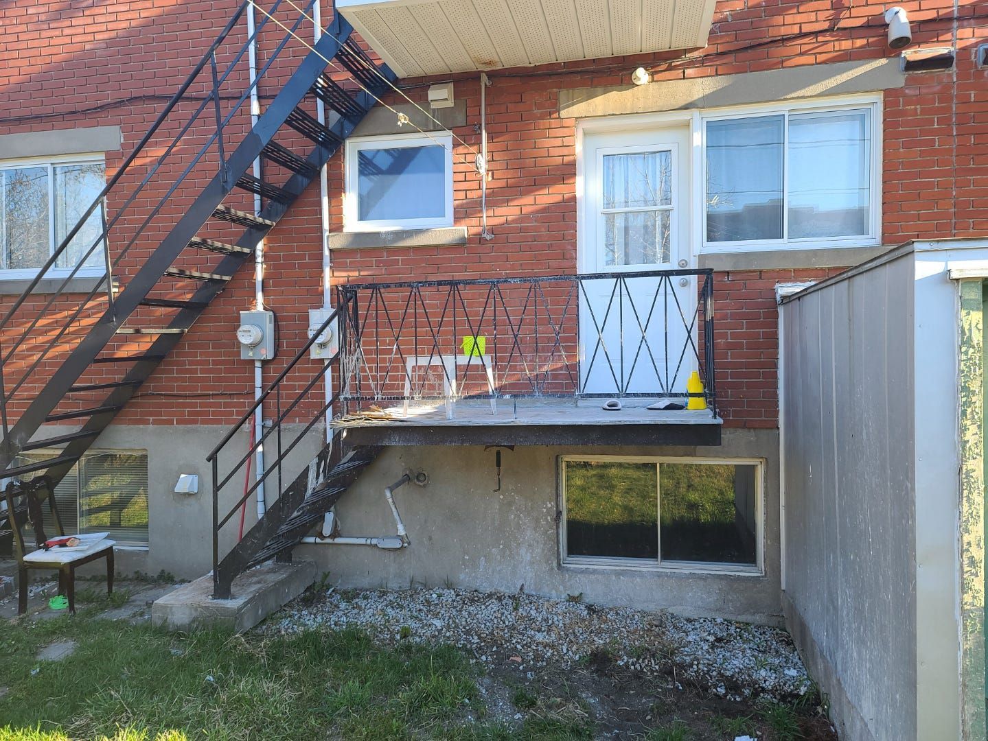Entrée d'appartement en briques avec escalier en métal noir, portes blanches et petit balcon donnant sur une cour gravillonnée
