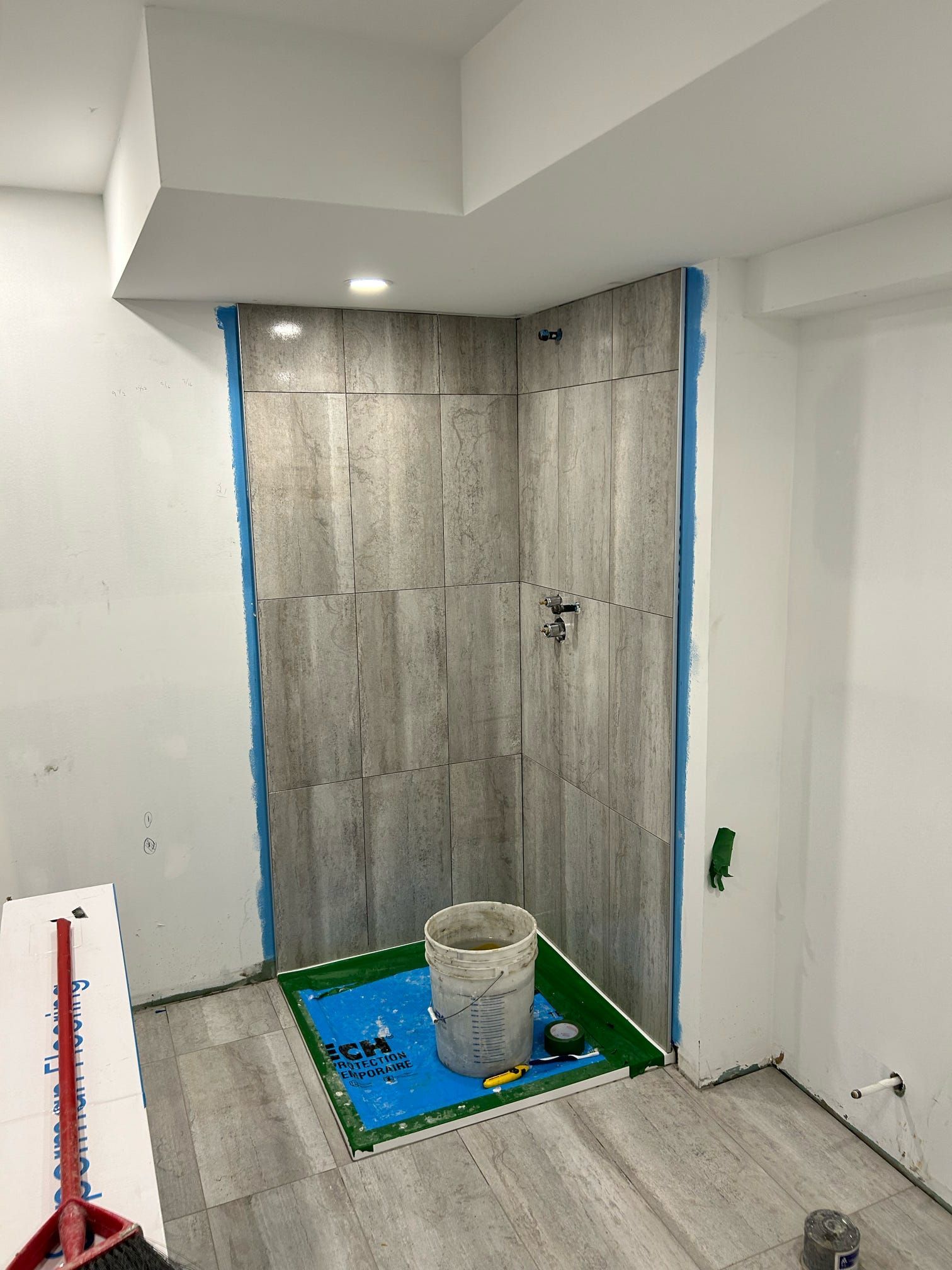 Petite salle de bain carrelée en cours de rénovation, avec des carreaux muraux gris, du matériel de peinture et un seau sur un tapis bleu.