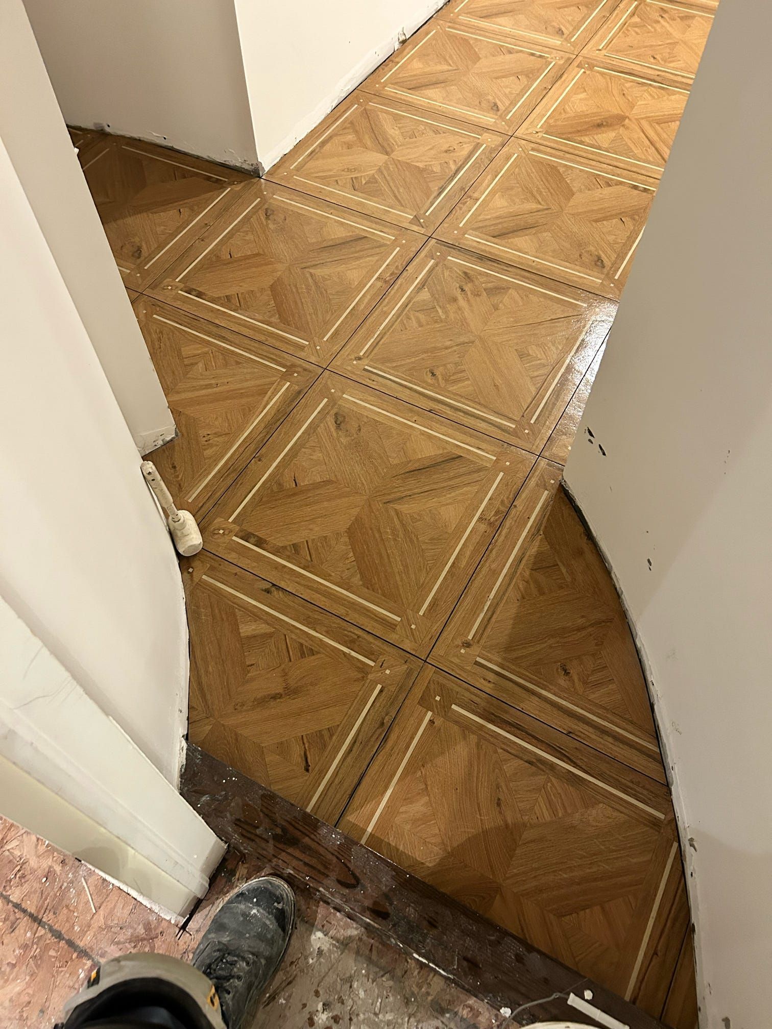 Couloir avec sol carrelé beige à motifs, vu entre des murs blancs et une porte, avec une chaussure en bas à gauche