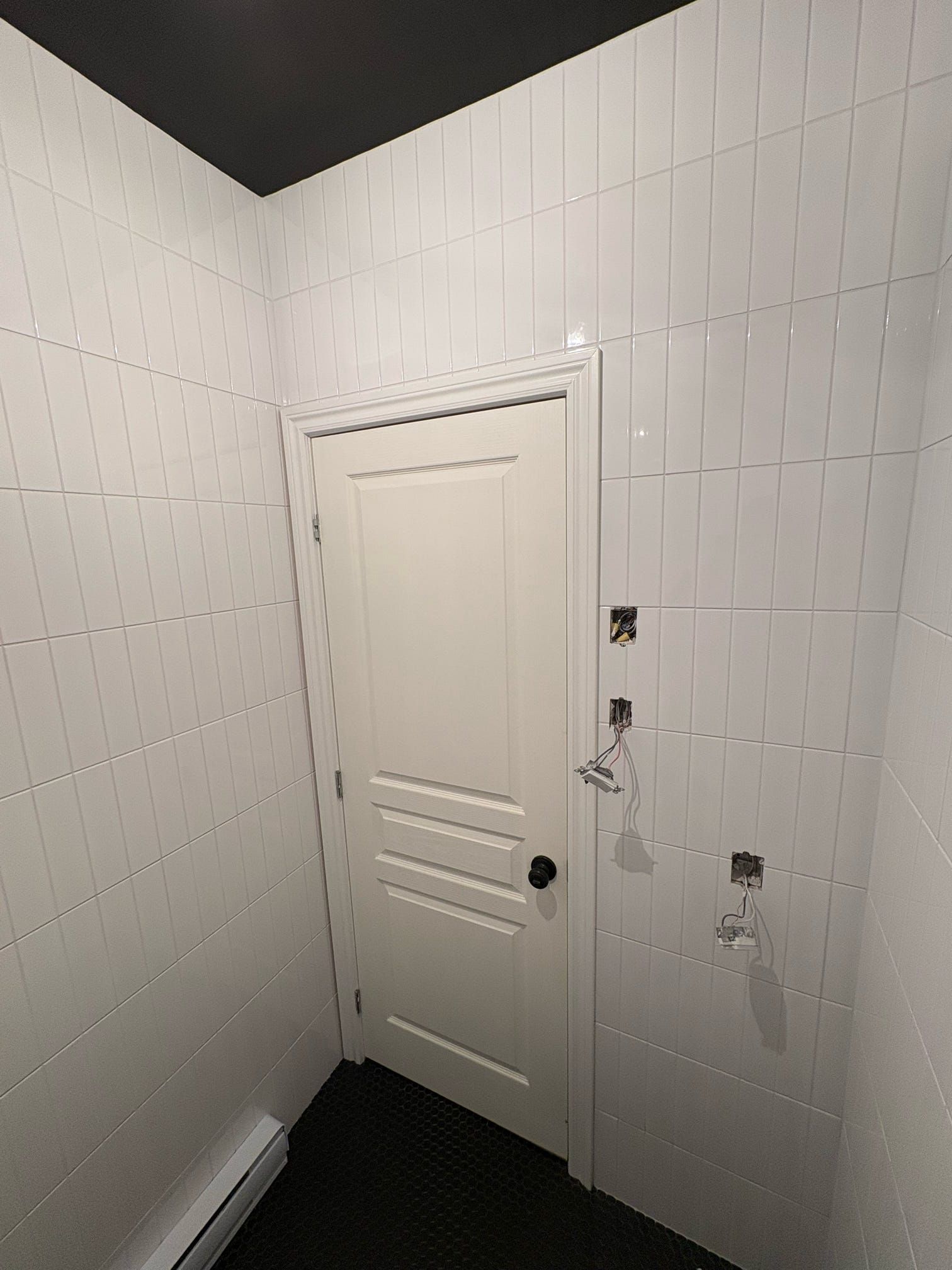 Petite pièce blanche aux murs lambrissés et à la porte fermée, avec des câbles apparents sur le mur de droite.