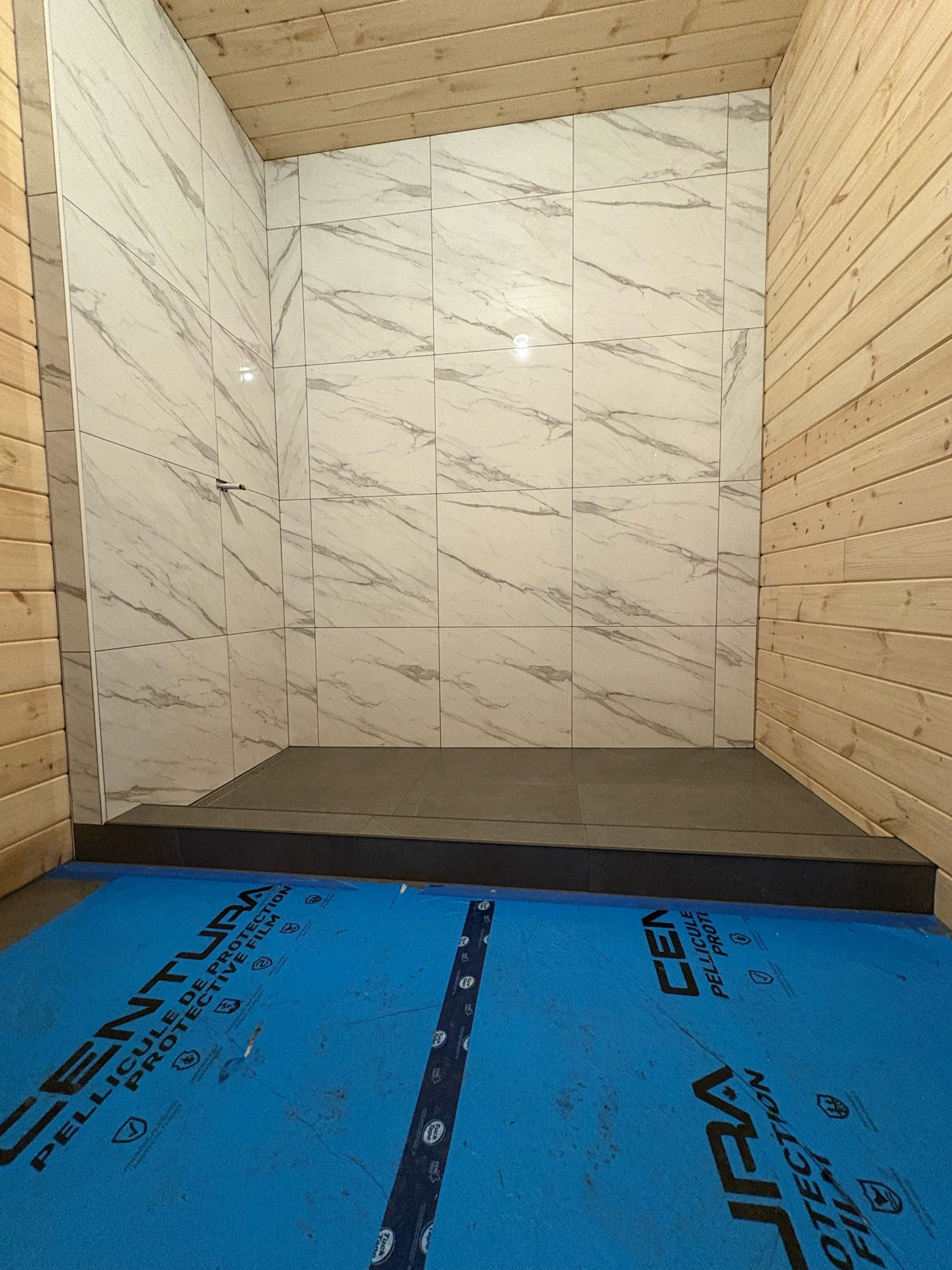 Alcôve de douche partiellement carrelée avec murs imitation marbre, panneaux latéraux en bois et protection de sol bleue
