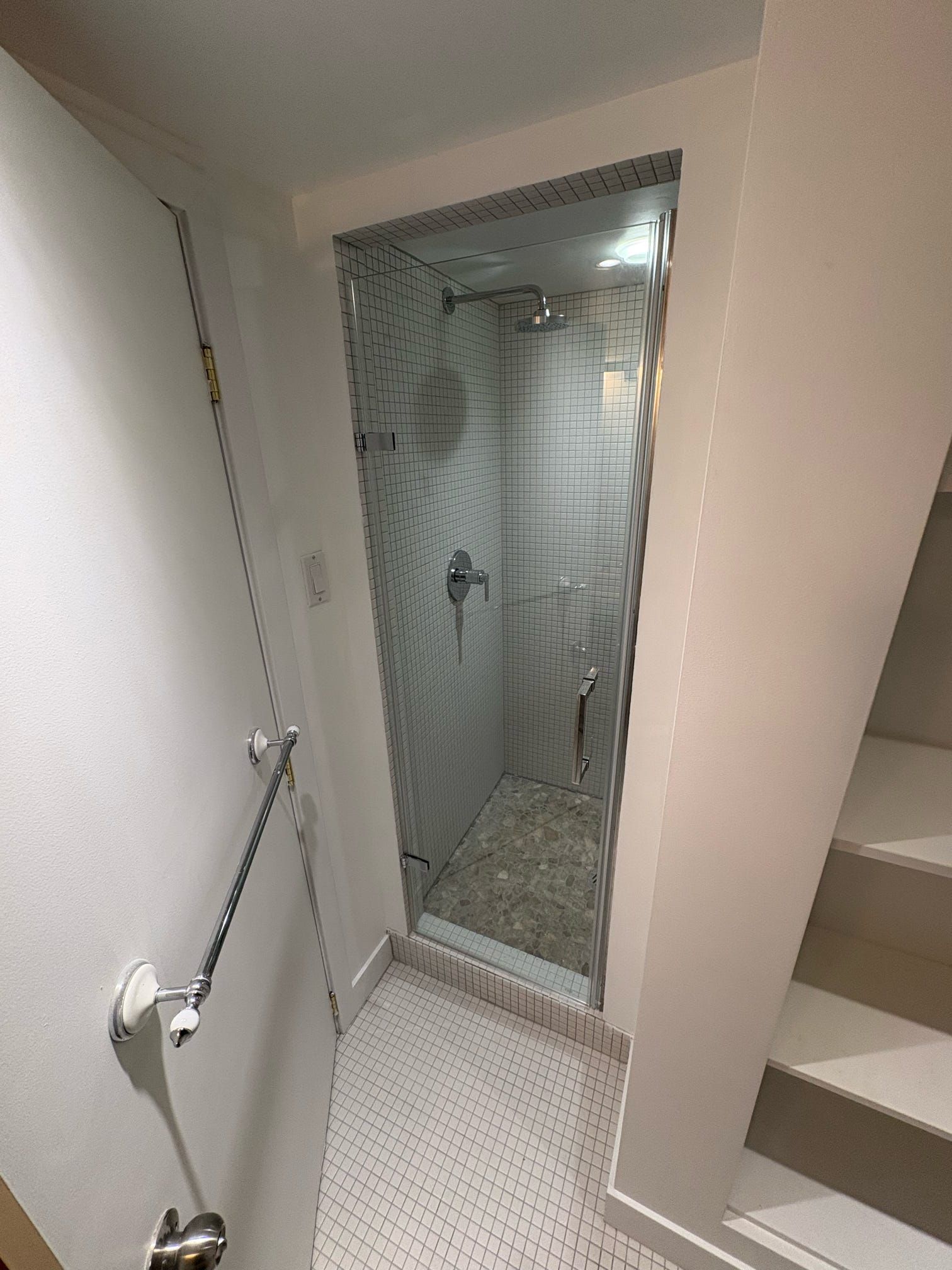 Salle de bain étroite avec cabine de douche vitrée, sol carrelé blanc et barre d'appui murale