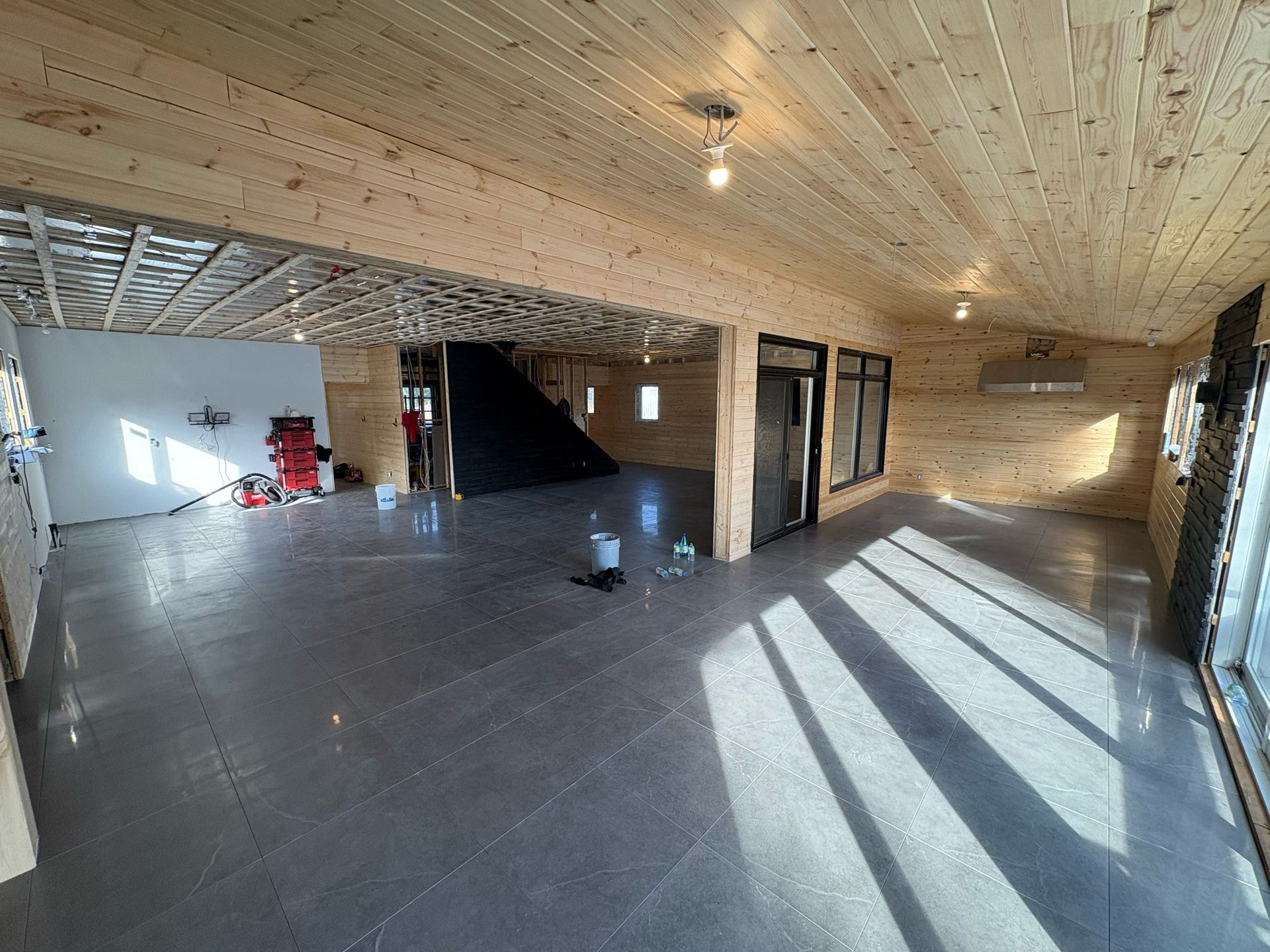 Pièce moderne spacieuse et vide, avec plafond en bois, carrelage gris au sol et lumière naturelle inondant les pièces grâce à de grandes fenêtres.