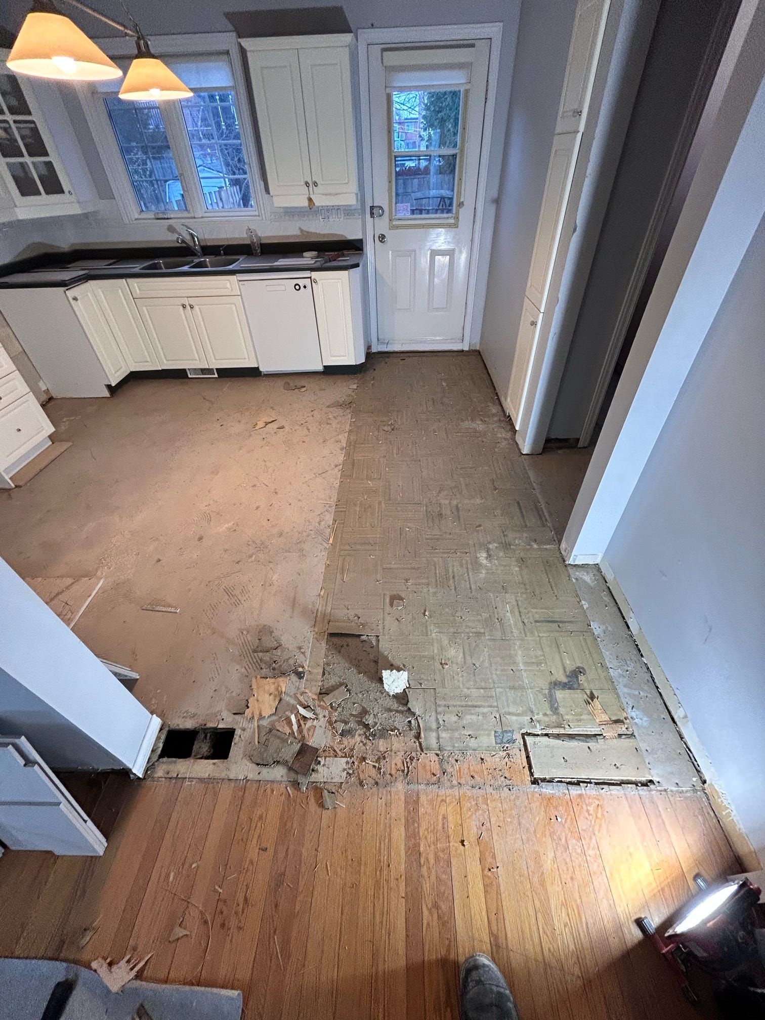 Sol de cuisine endommagé, moquette manquante, sous-plancher apparent et placards blancs près d'une porte arrière
