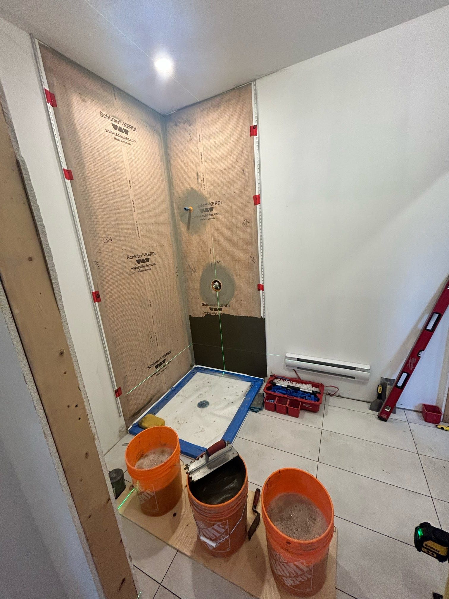 Petite salle de bain en rénovation avec parois de douche apparentes, seaux, outils et carrelage au sol.