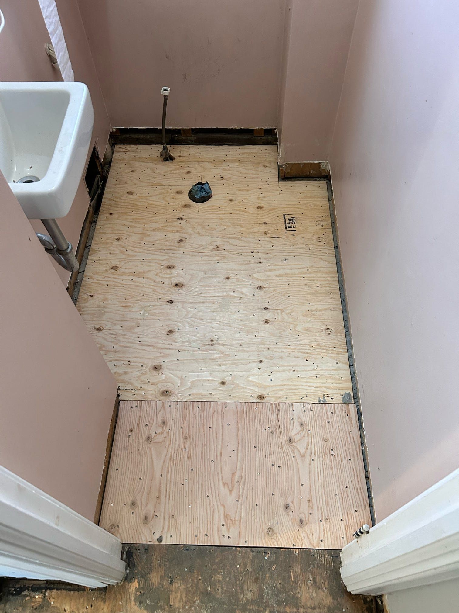 Salle de bain étroite et inachevée avec plancher apparent, un lavabo sur la gauche et une ouverture d'évacuation près du fond.