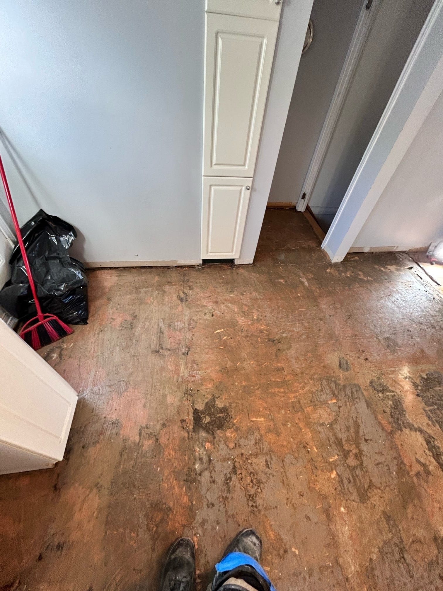 Couloir étroit au sol marron usé, portes blanches, un sac-poubelle et un balai appuyé contre le mur.