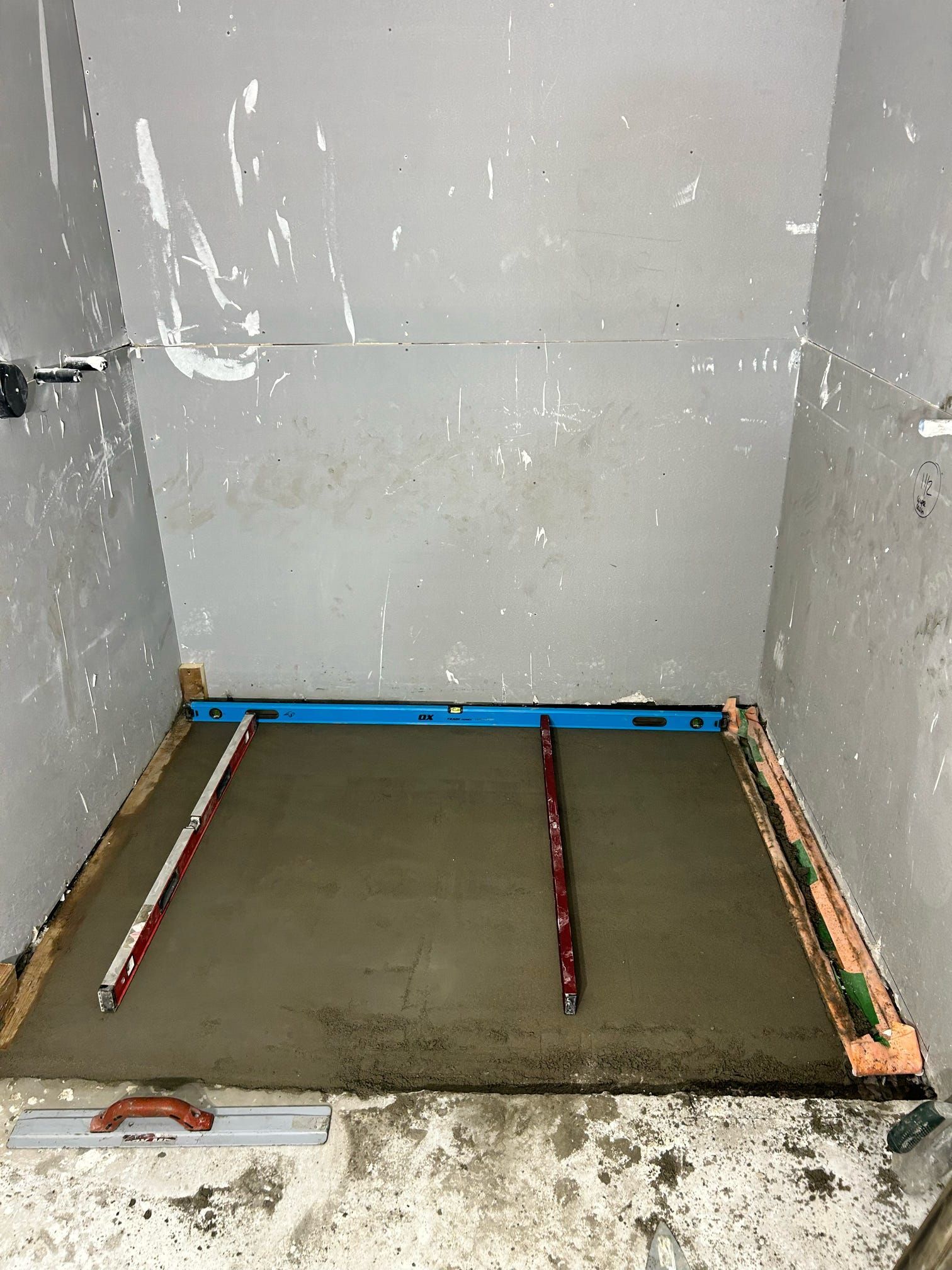 Sol de douche non fini avec receveur en ciment gris et tuyaux apparents contre les murs carrelés