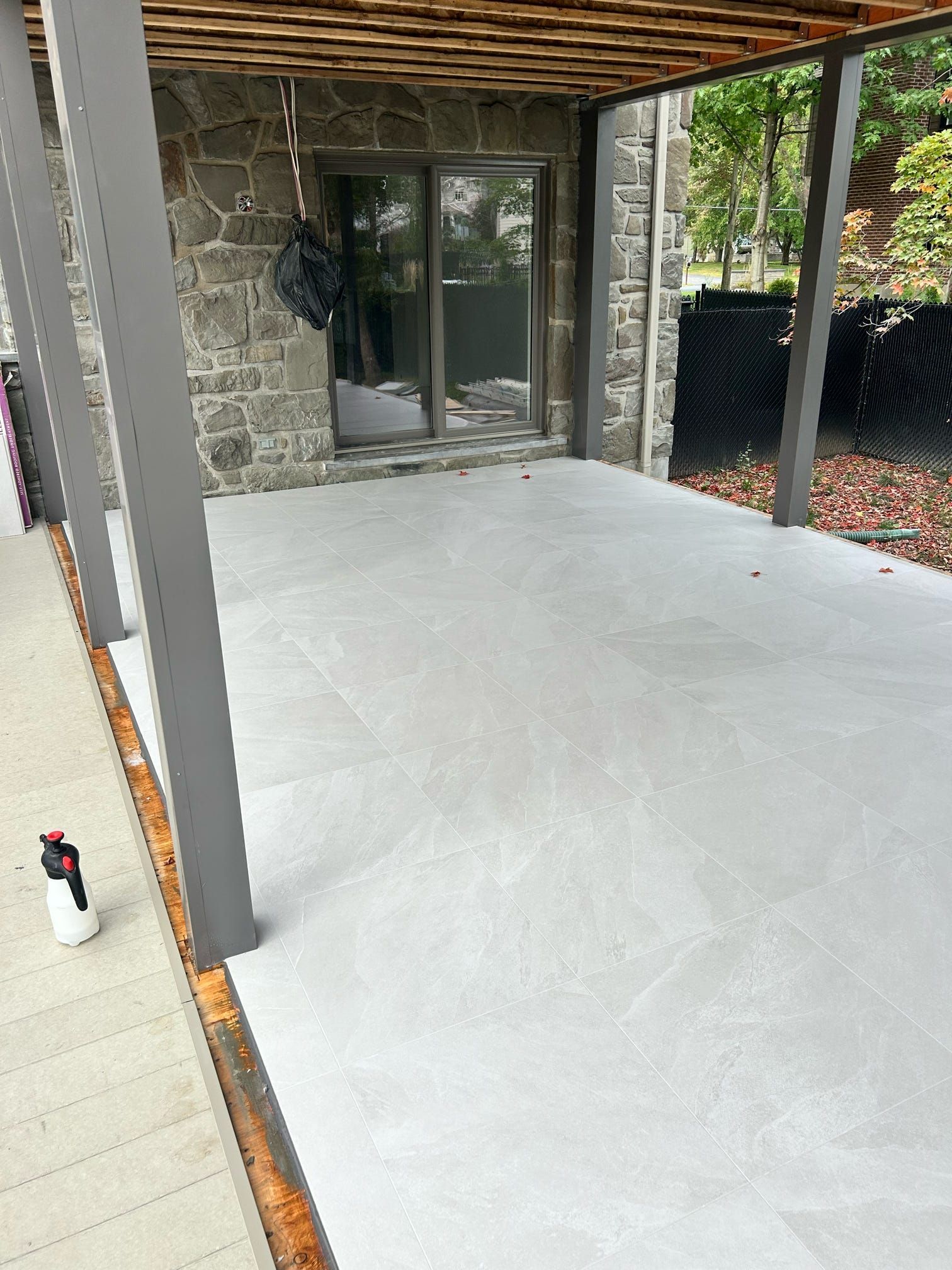 Patio couvert avec sol lisse gris clair, mur en pierre, porte coulissante en verre et plafond en pergola de bois.