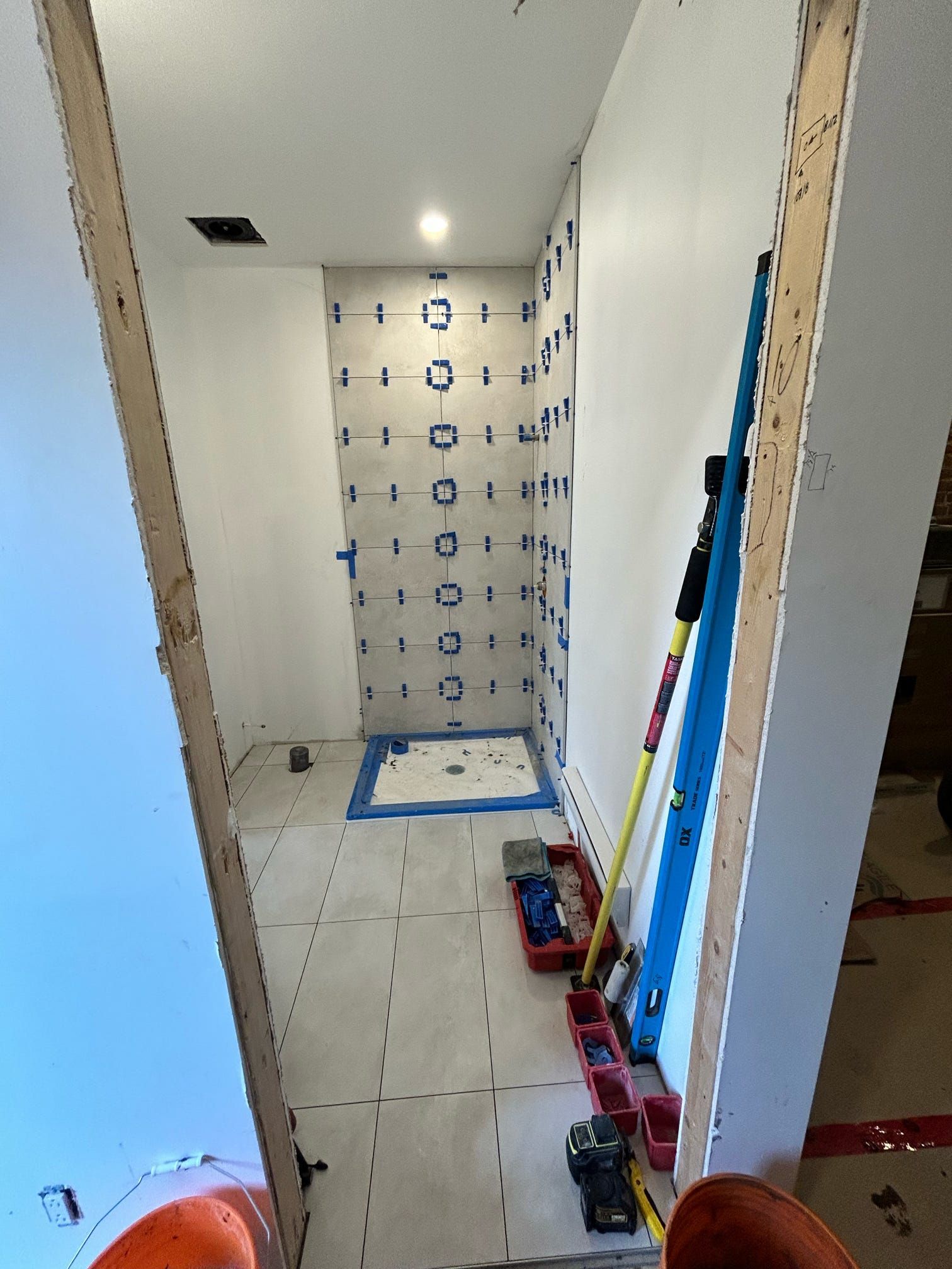 Couloir étroit et inachevé avec sol carrelé, marquages ​​au ruban adhésif bleu et outils appuyés contre le mur.