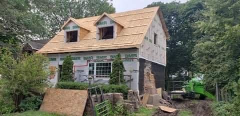 Petite maison en rénovation avec un toit beige, des murs inachevés et des arbres tout autour.